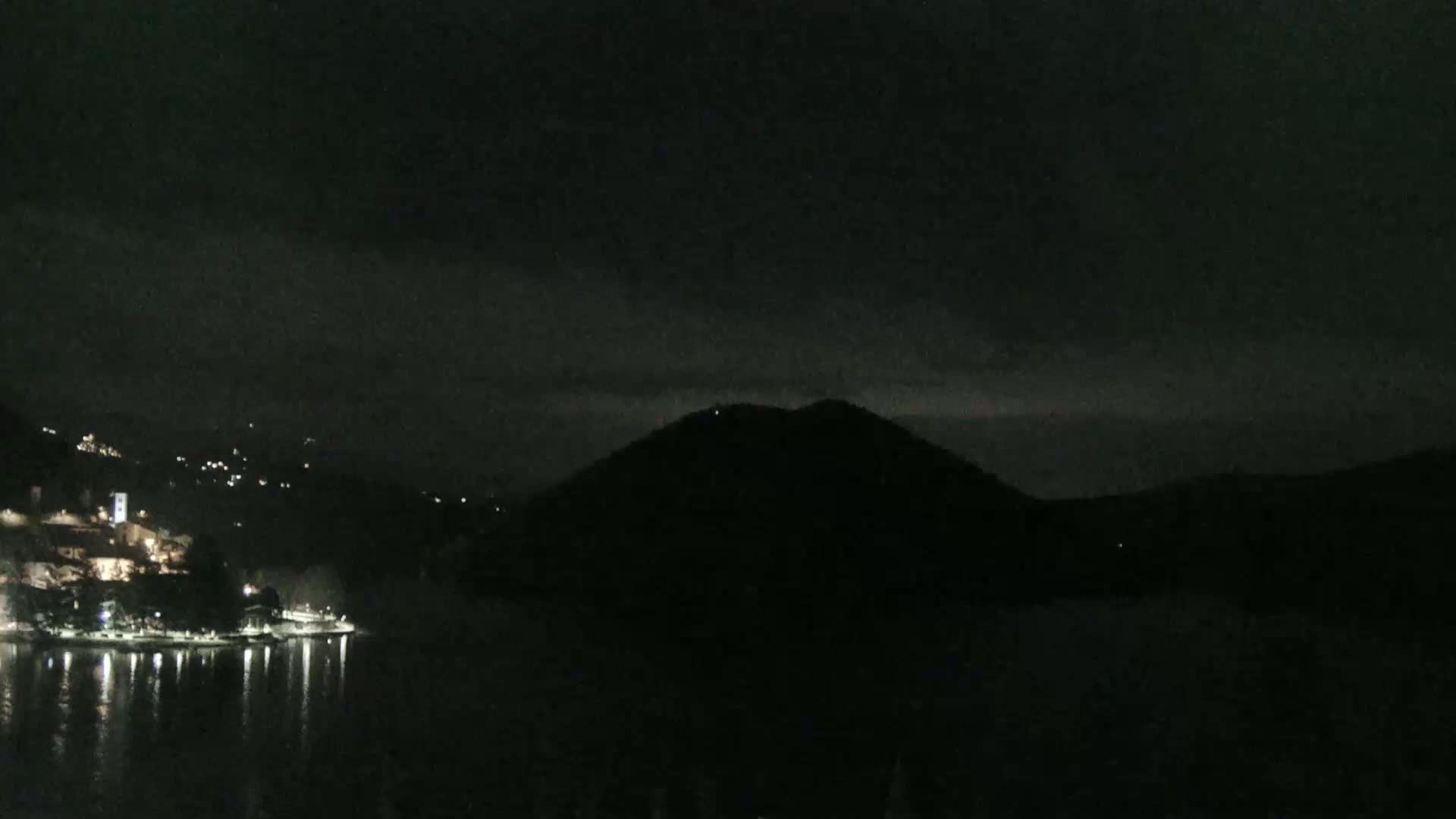 A nighttime view of a dark, hilly landscape with a body of water reflecting scattered lights from a town on the shore under a mostly cloudy sky.