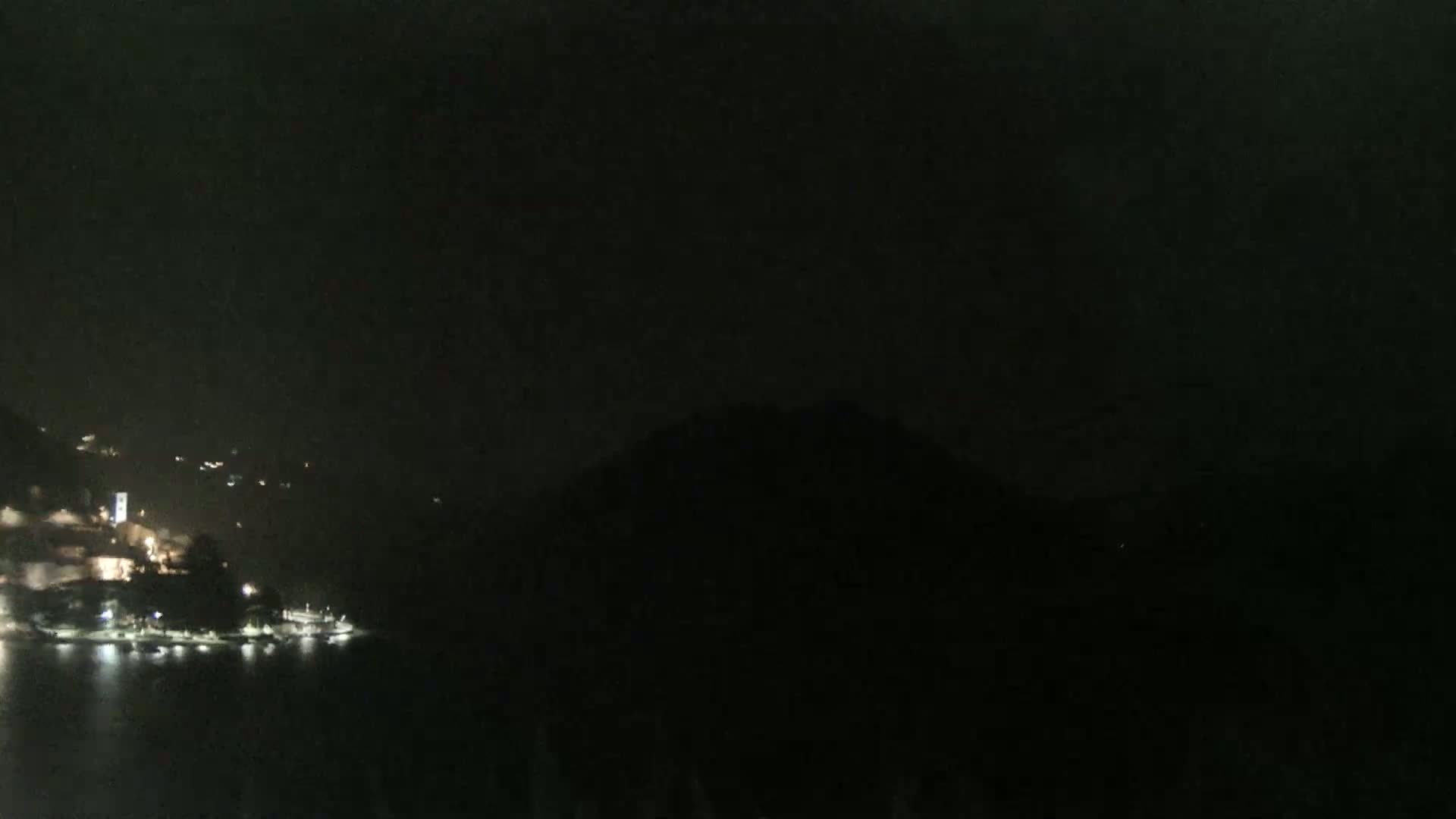 A nighttime view of a dark body of water reflecting the lights of a town nestled against a hillside under a dark, cloudy sky.
