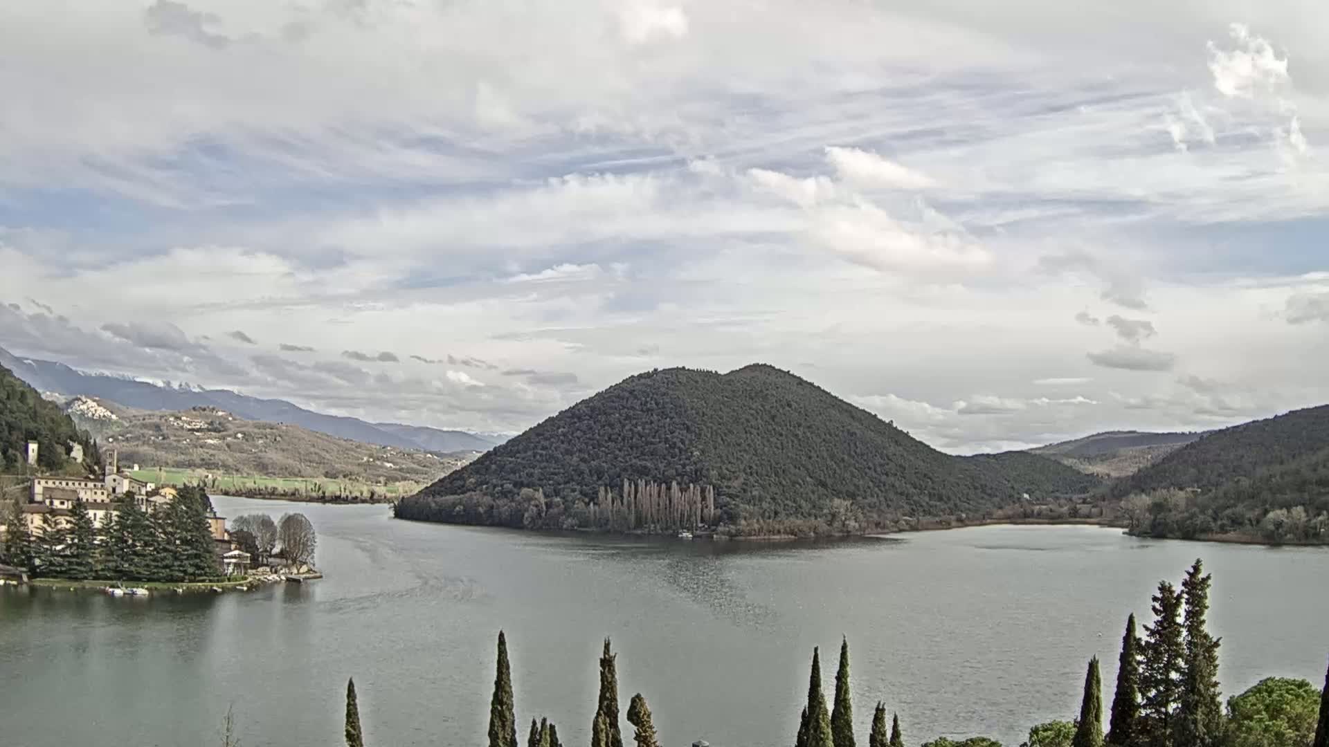 A lake with a village and a tree-covered hill is seen under a mostly cloudy sky.