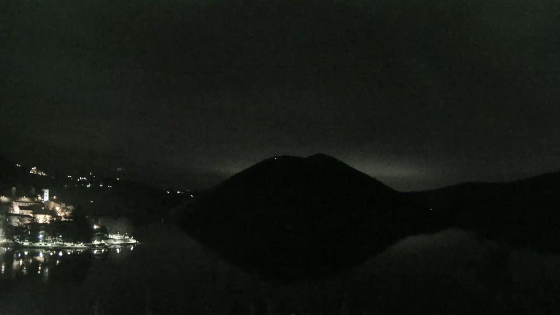 A dark, nighttime scene shows a body of water reflecting the faint lights of a town nestled in the hills, with a large, silhouetted hill in the center.