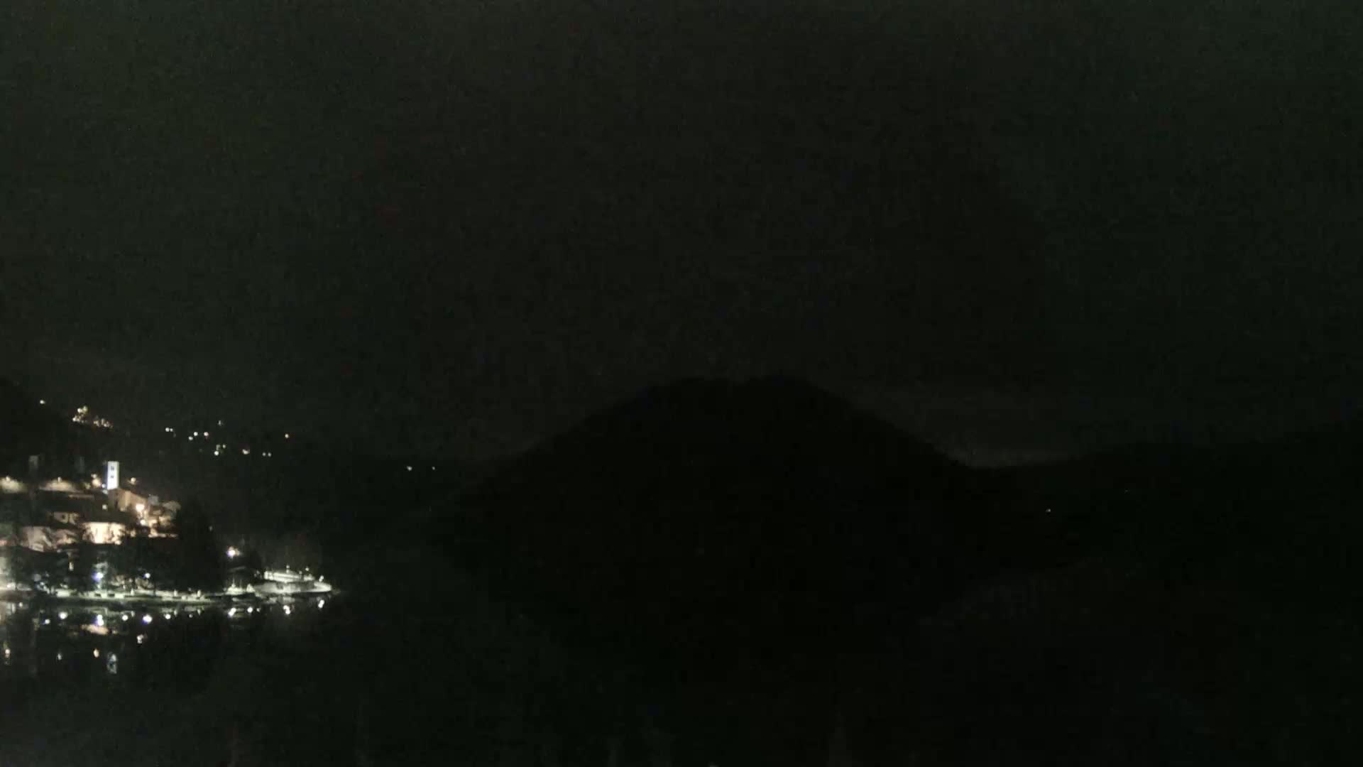 A dark night scene shows a dimly lit town on a body of water with a large, dark hill or mountain in the background.