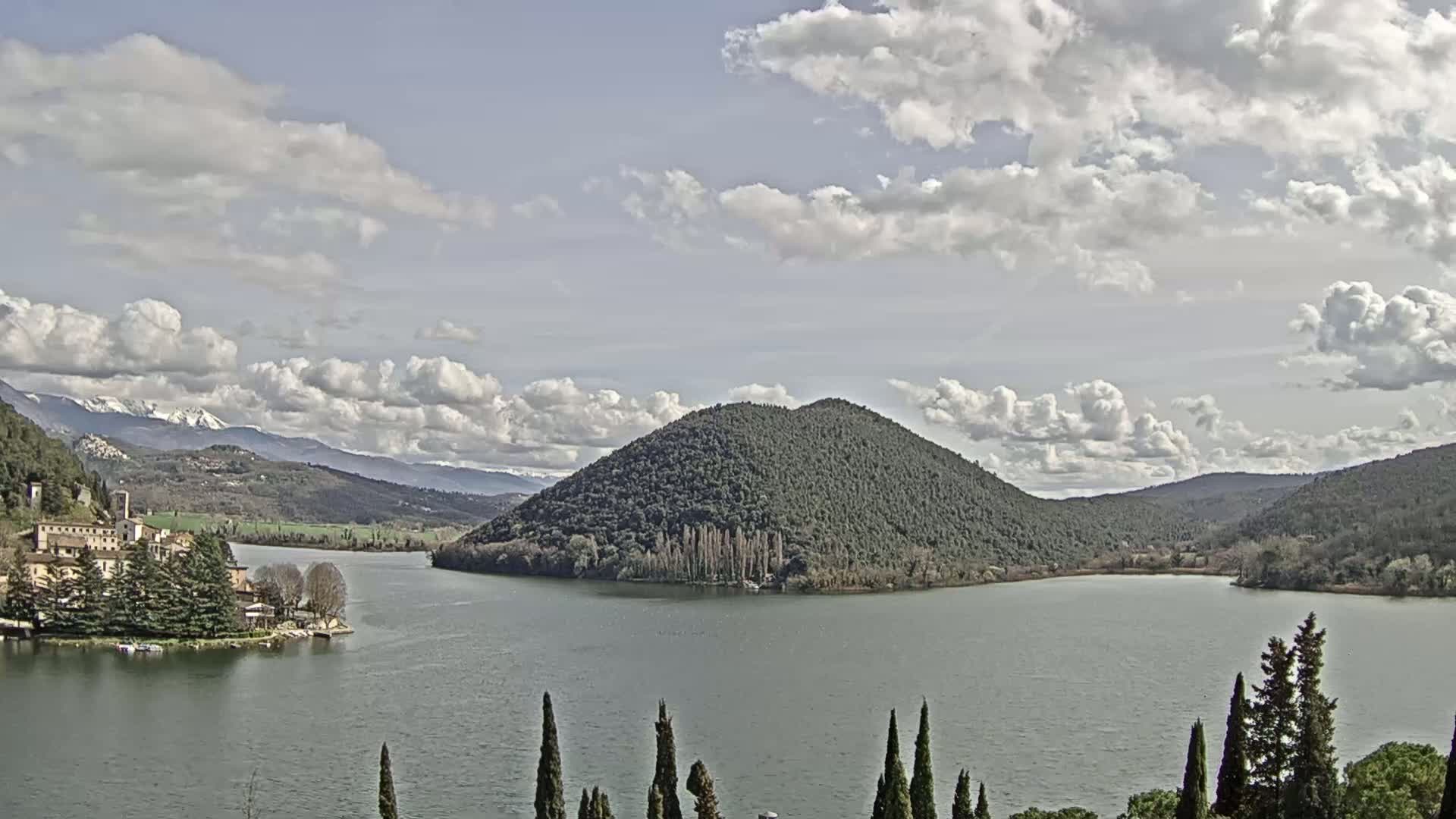 A partly cloudy day reveals a lake with a tree-covered island, a village nestled on the shore, and snow-capped mountains in the distance.
