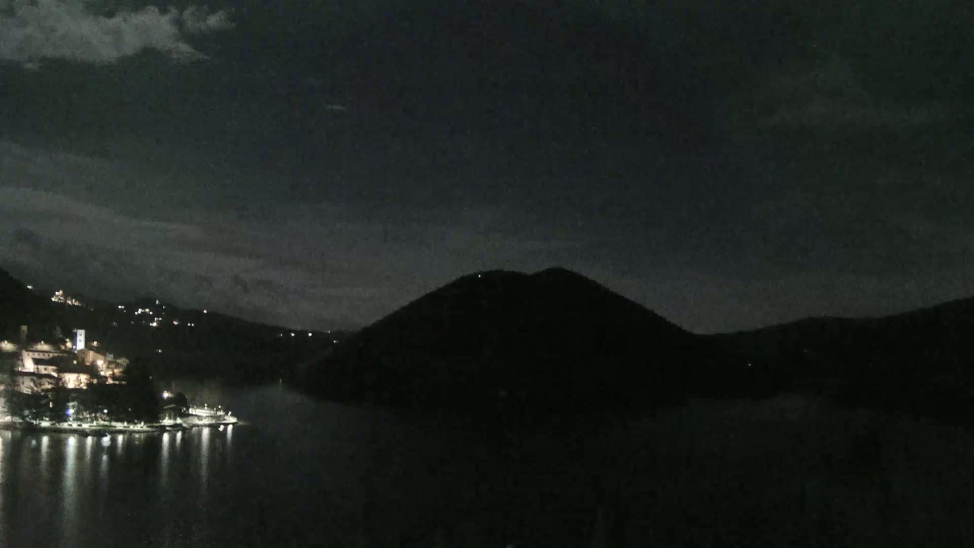 A nighttime view shows a dark mountain across a body of water reflecting the lights of a small, illuminated town on the opposite shore under a mostly cloudy sky.