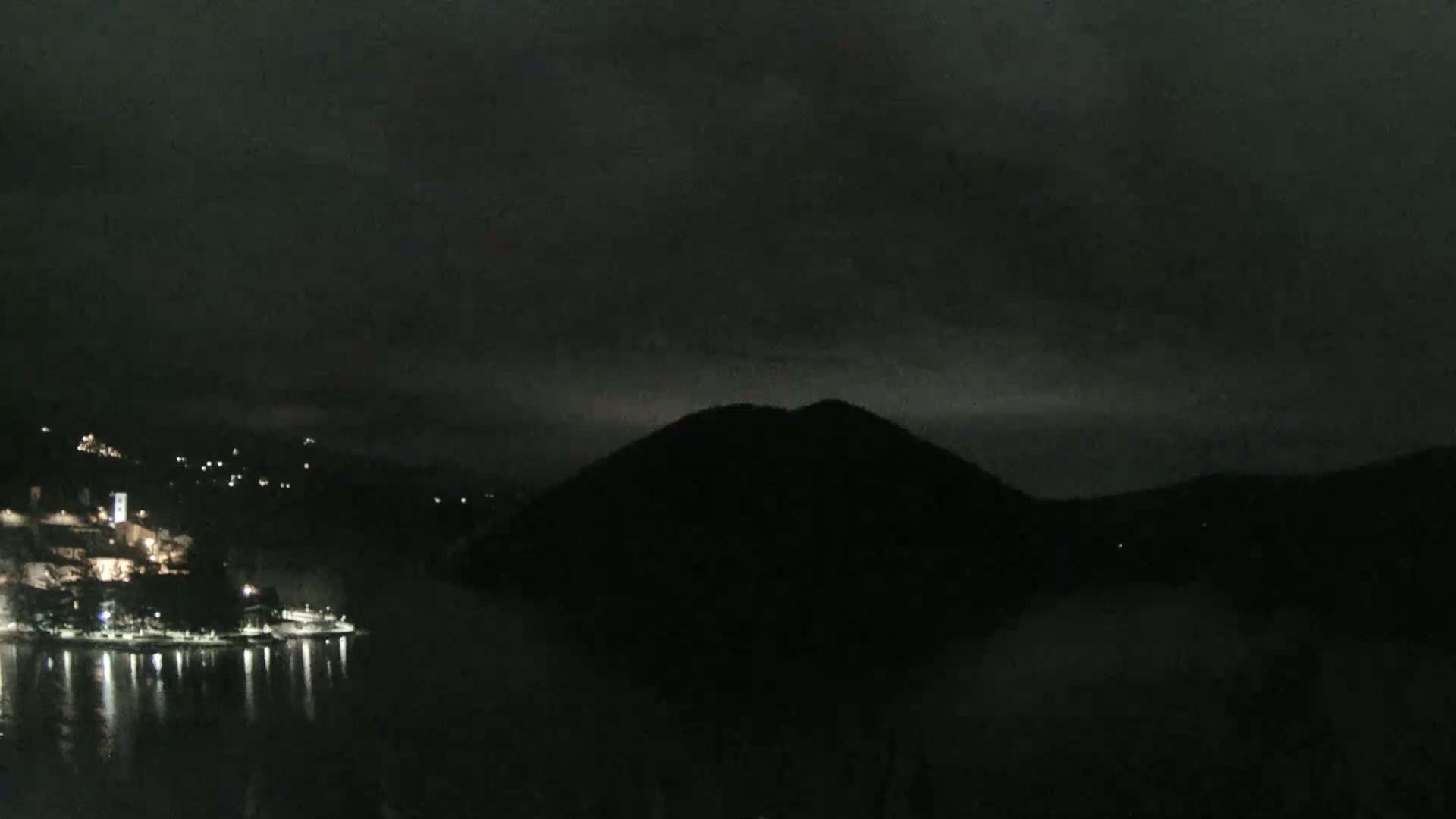 A nighttime view of a dark, hilly landscape with a body of water reflecting the lights of a small town nestled along its shore under a cloudy sky.