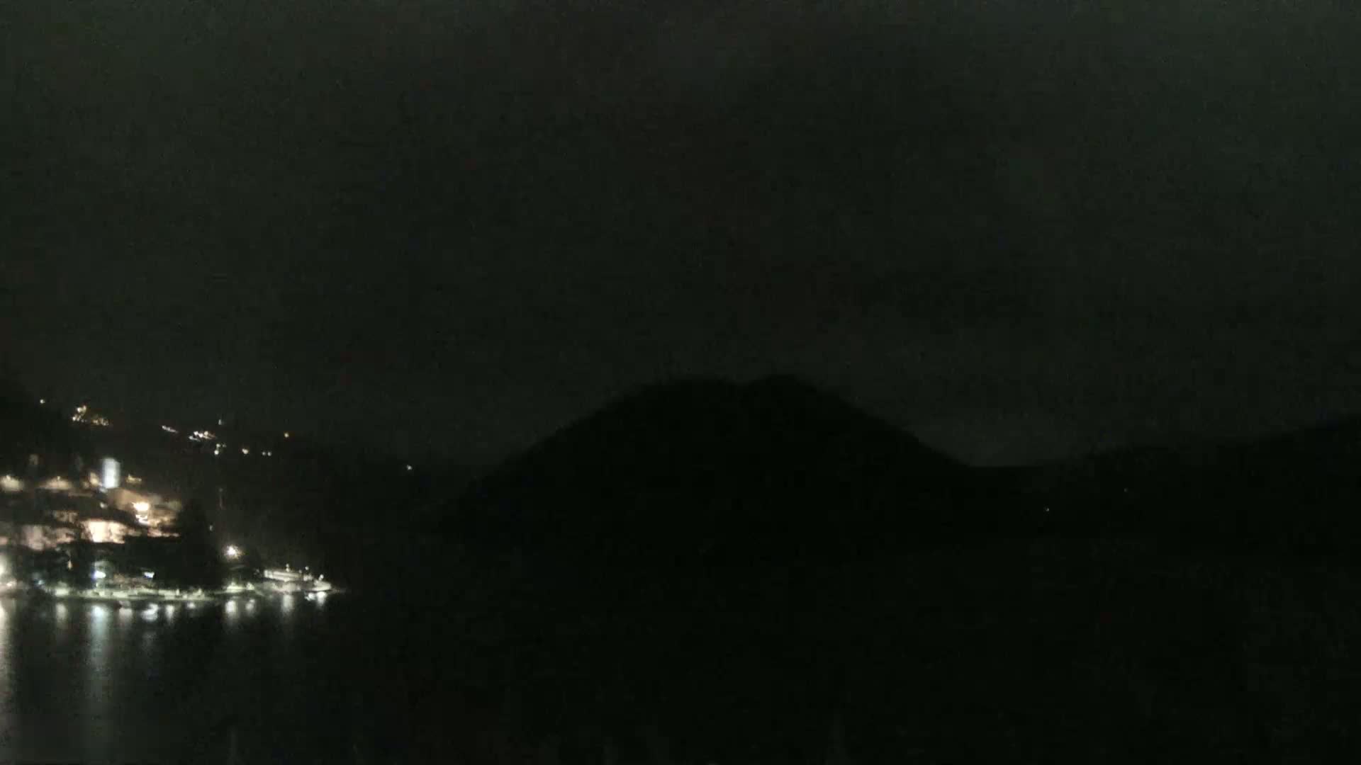 A nighttime view of a dark, calm body of water with a hill silhouetted against a cloudy sky, and faintly lit buildings on the far shore.