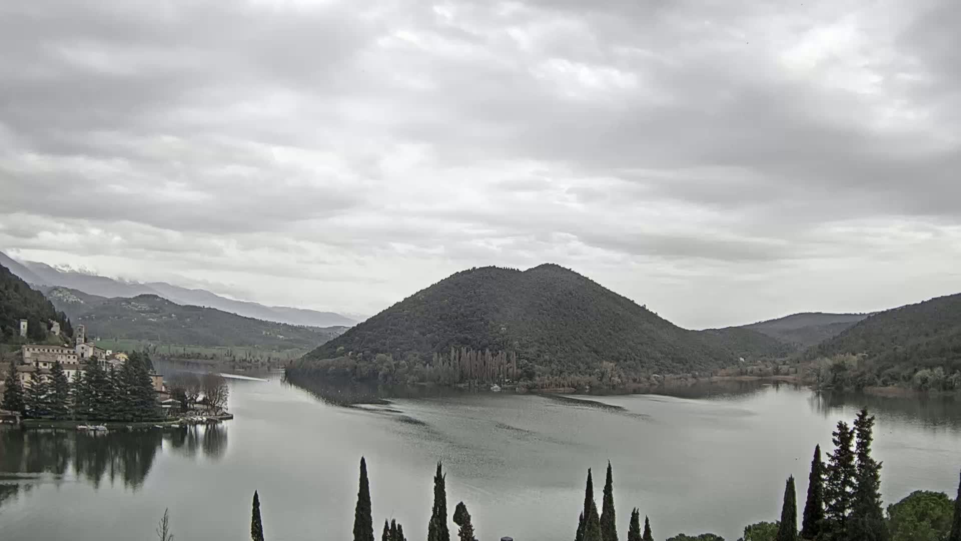 A lake surrounded by dark green hills and a village under an overcast sky.