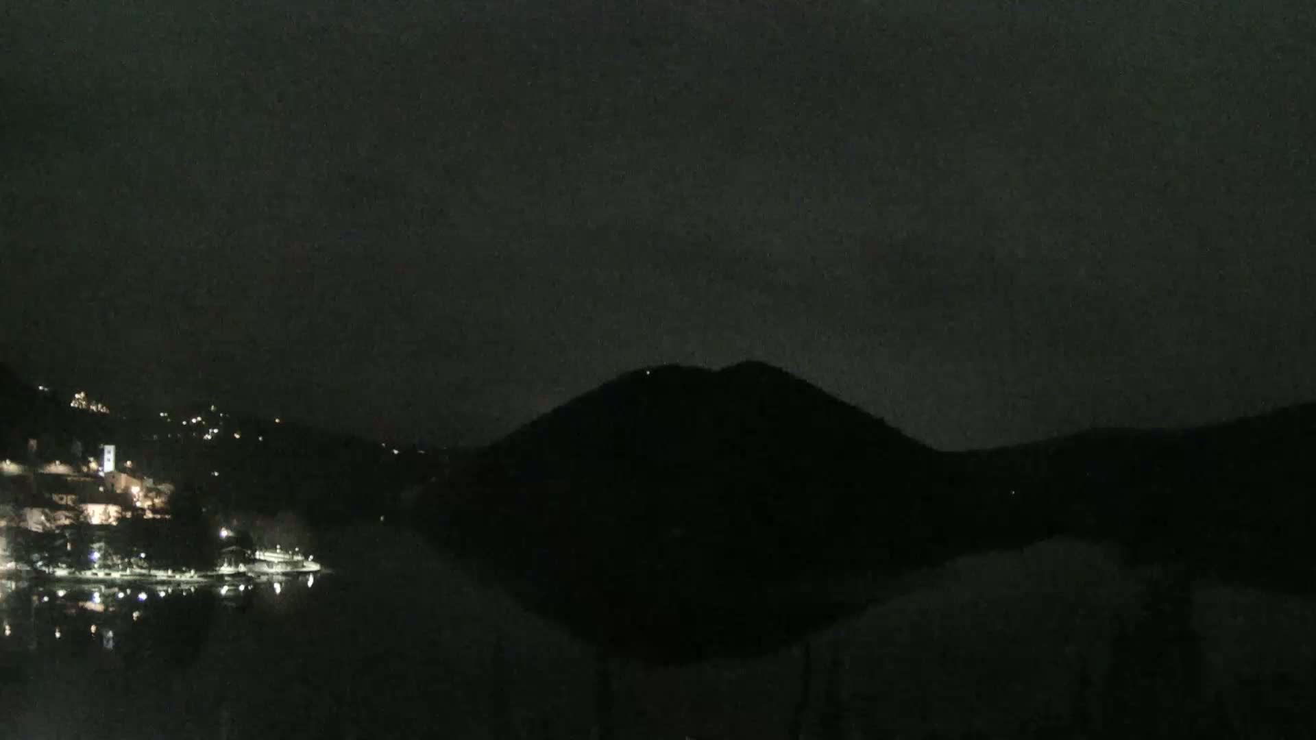 A dark, nighttime scene shows a body of water reflecting the lights of a town nestled on a hillside, with a large, silhouetted hill in the background under a cloudy sky.