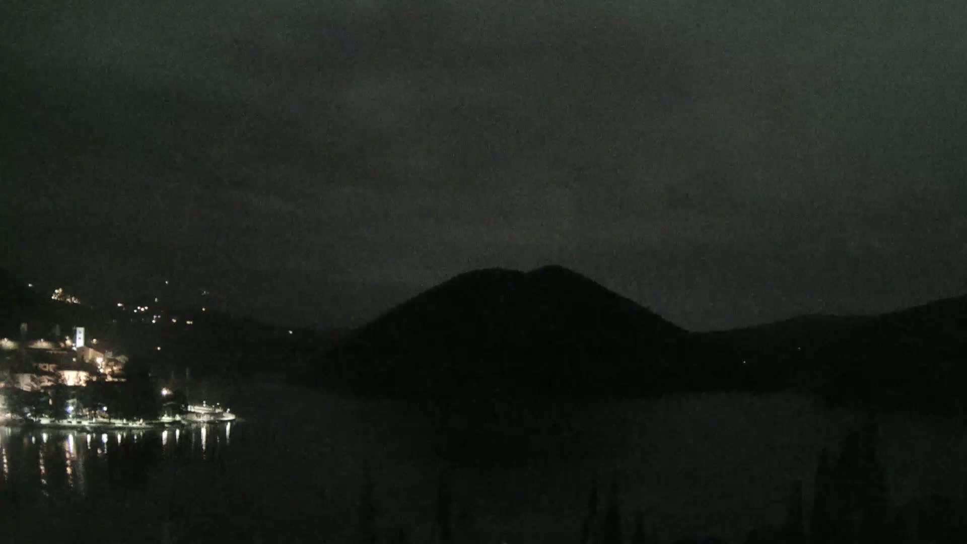A nighttime view shows a dark, silhouetted hill across a body of water reflecting scattered lights from a small, illuminated town on the opposite shore under a cloudy sky.