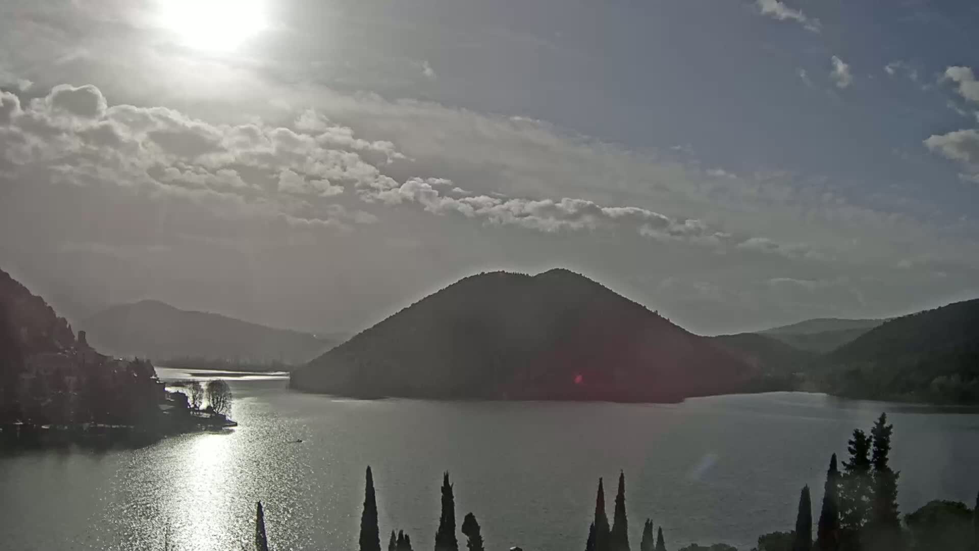 A partly cloudy day reveals a sunlit lake surrounded by dark, rolling hills.