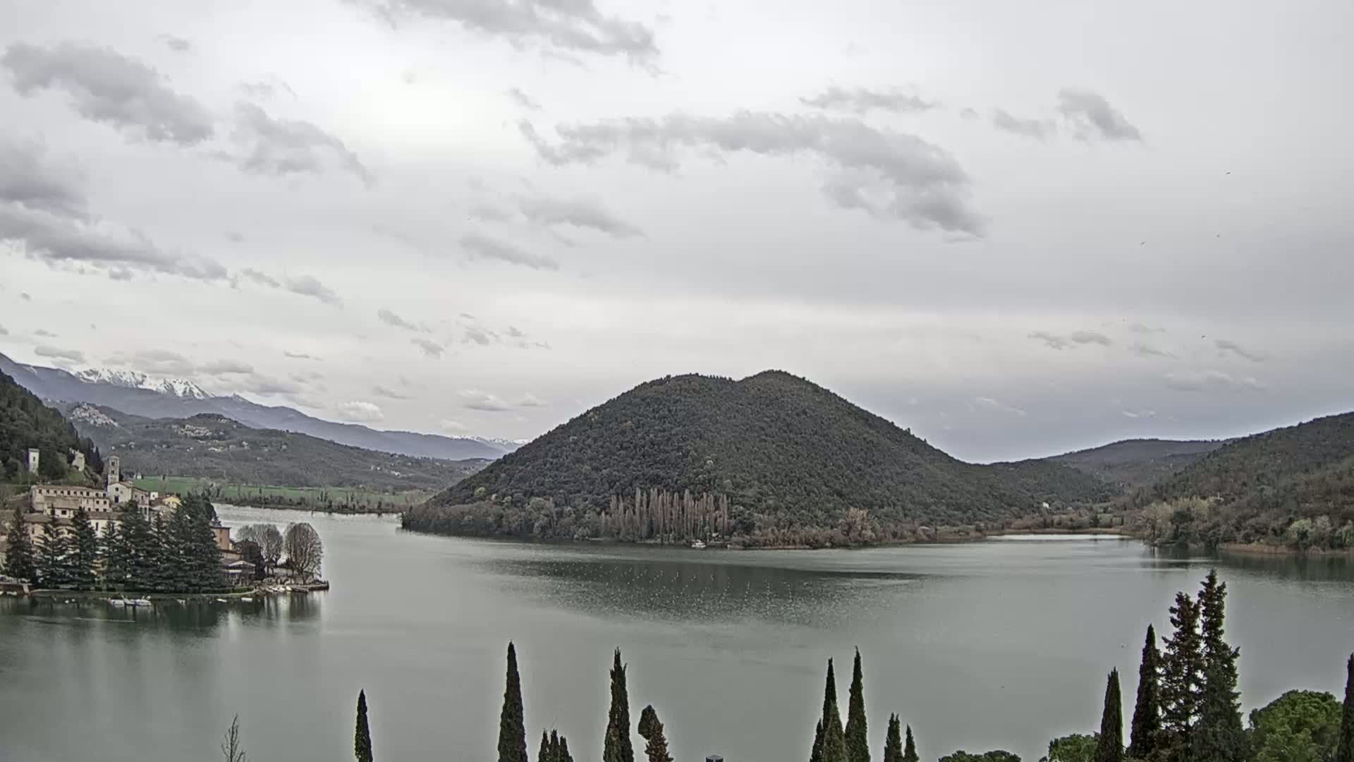 A lake is surrounded by hills and mountains under a cloudy sky, with a village nestled on the lakeshore and snow-capped mountains in the distance.