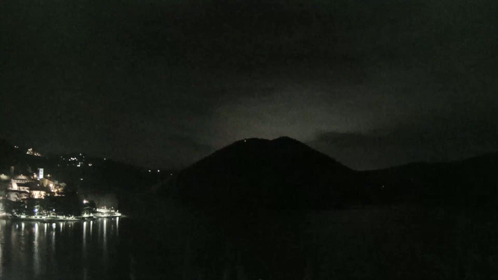 A dark, nighttime scene shows a body of water reflecting the lights of a town nestled at the base of a hill.