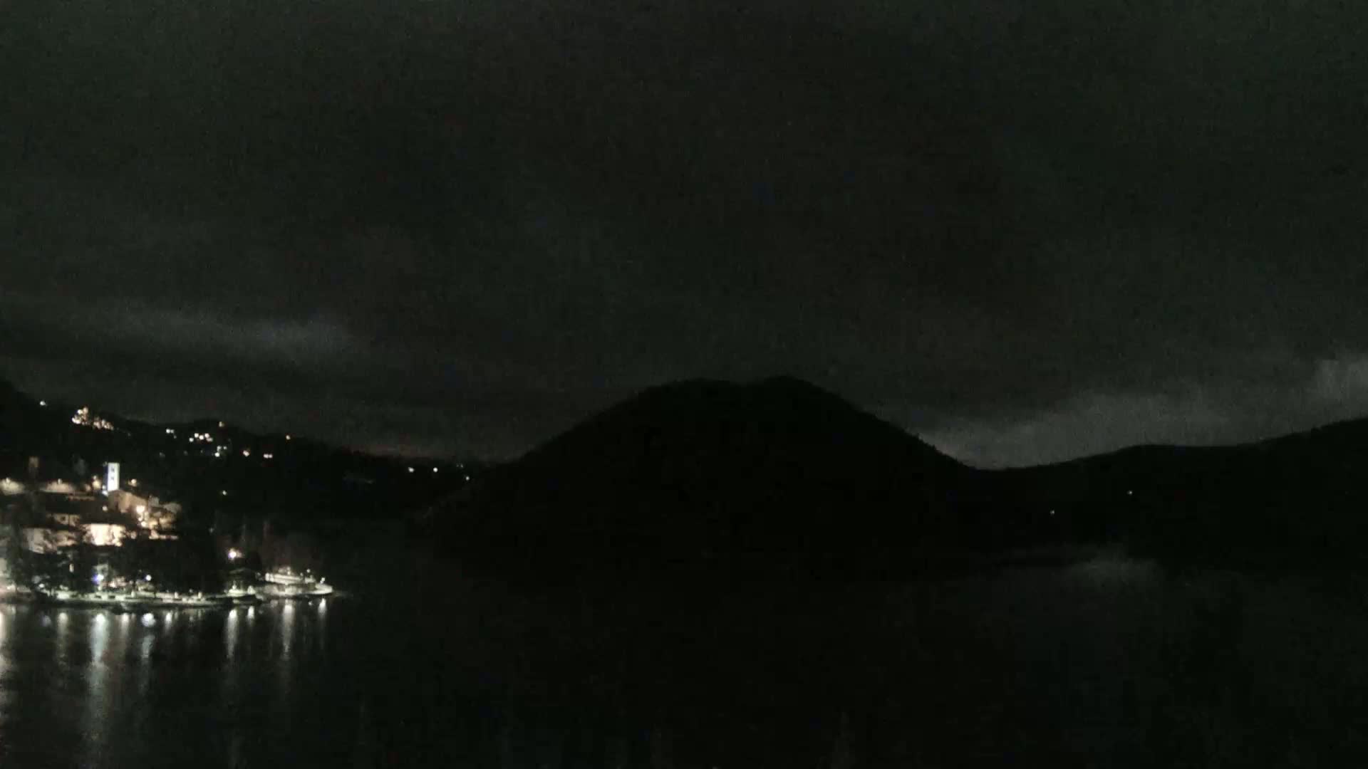 A dark nighttime scene shows a body of water reflecting the lights of a town nestled on a hillside, with a large, dark hill dominating the background under a mostly cloudy sky.