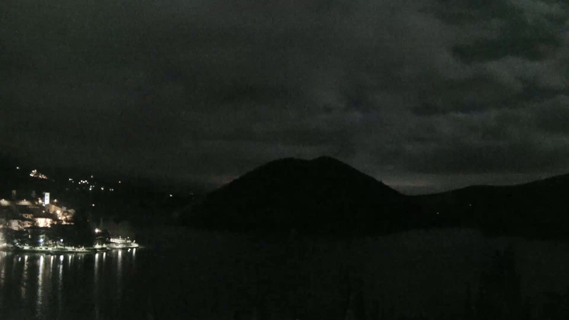 A dark, cloudy night scene shows a body of water reflecting the lights of a town nestled against a hill, with a larger, silhouetted hill in the background.