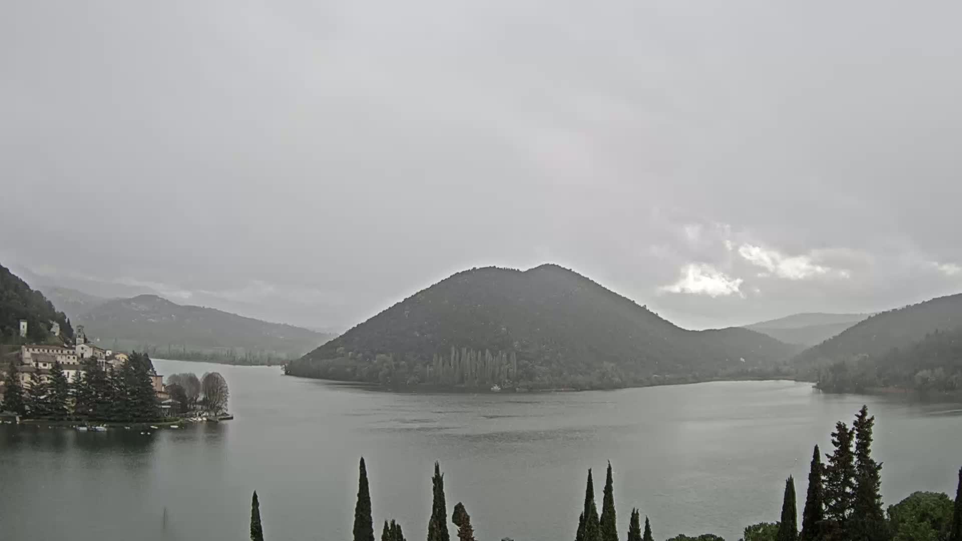 A lake is surrounded by hills and mountains under an overcast sky, with a small village visible on the left shore.