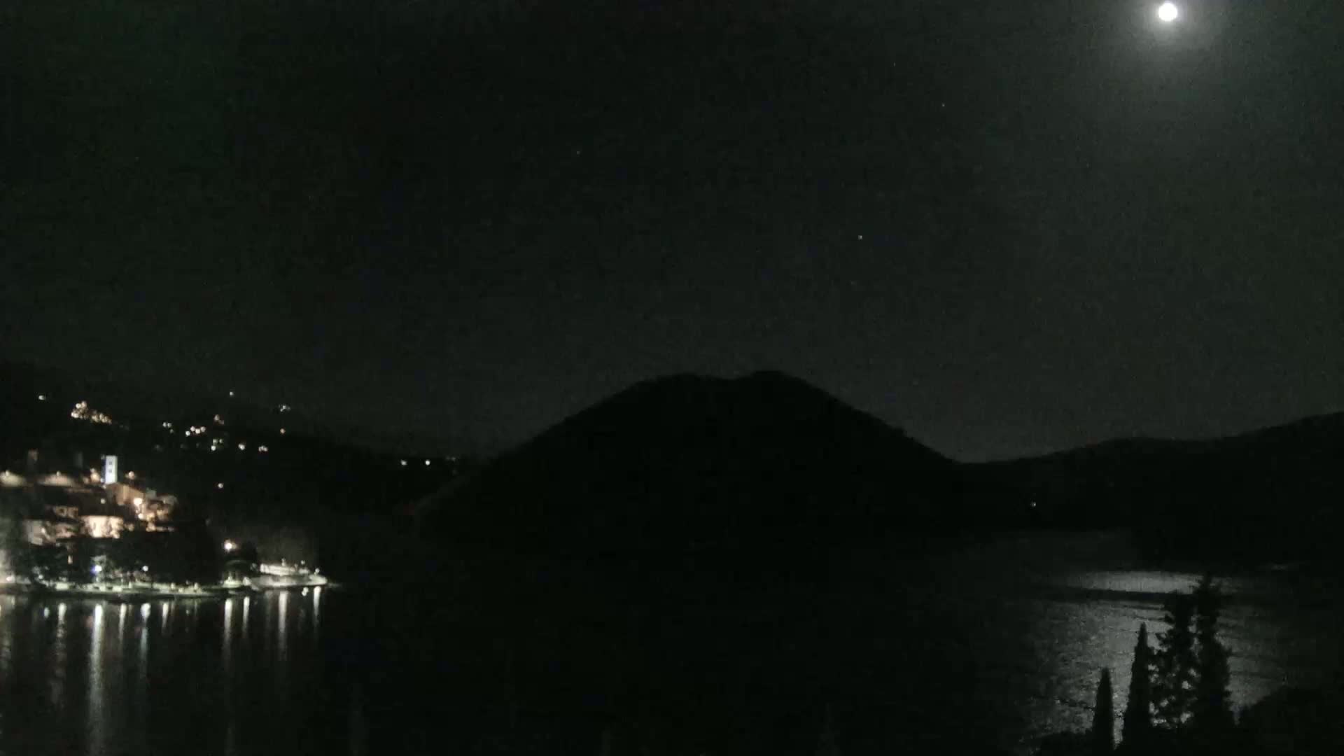 A moonlit night scene shows a body of water reflecting the light, a dark hill silhouetted against the sky, and a small town with lights visible on the opposite shore.