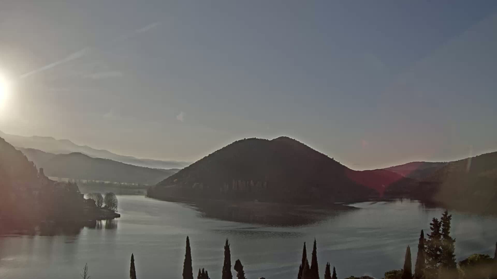 A calm lake is surrounded by hills and mountains under a clear, bright sky at sunrise.