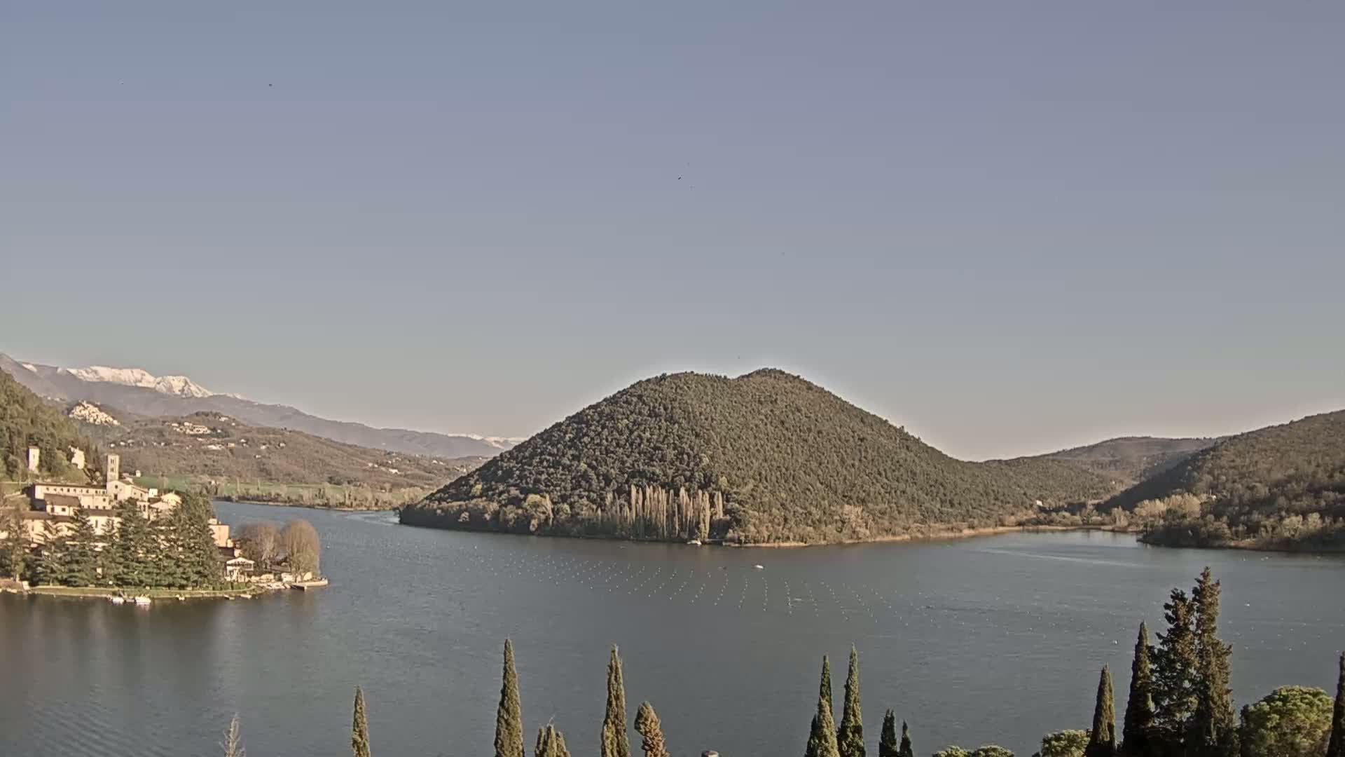 A lake is surrounded by hills and mountains, some of which are snow-capped, under a clear sky.