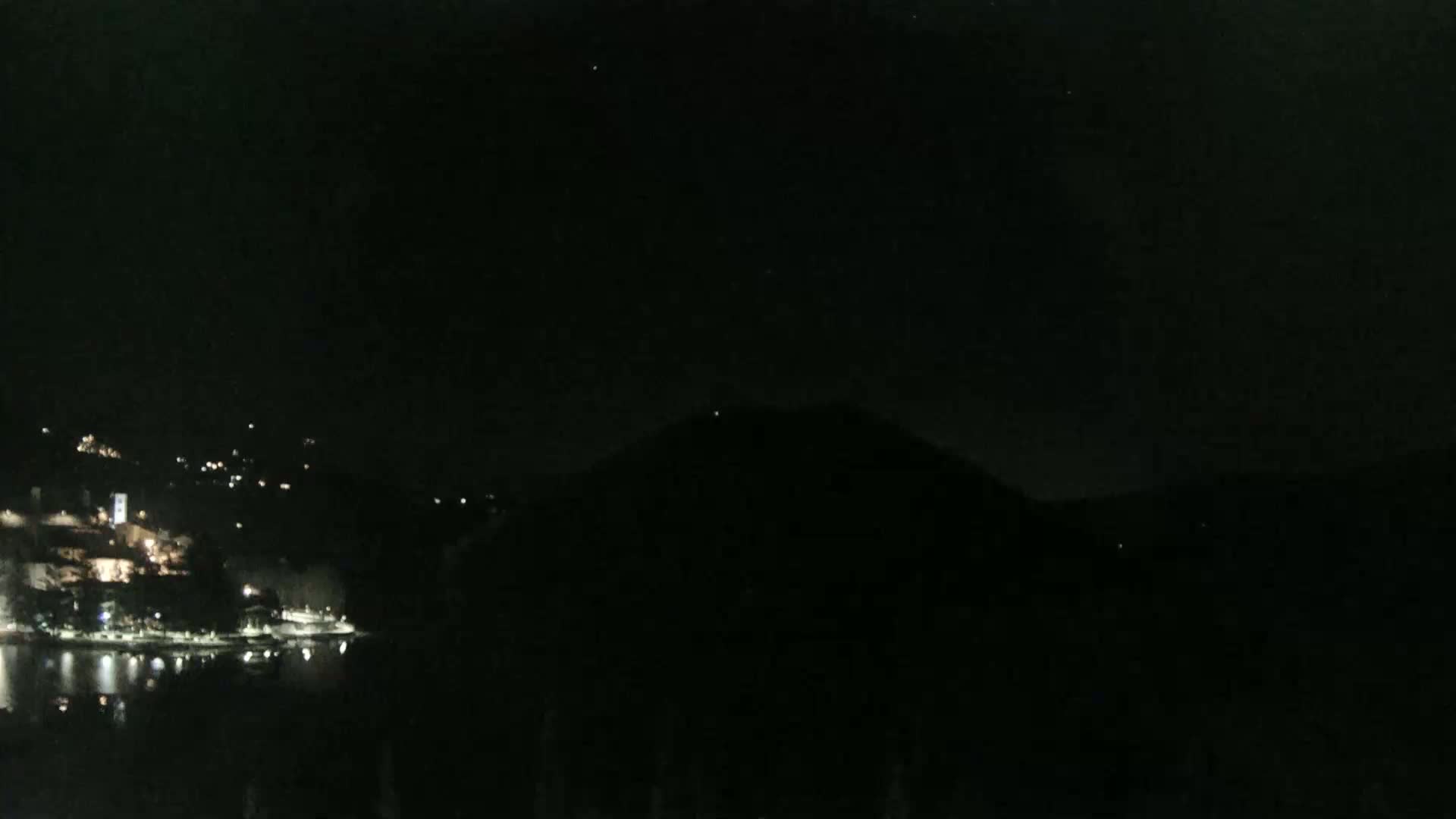 A nighttime view shows a dark, silhouetted hill overlooking a body of water with a town's lights reflecting on the water's surface.