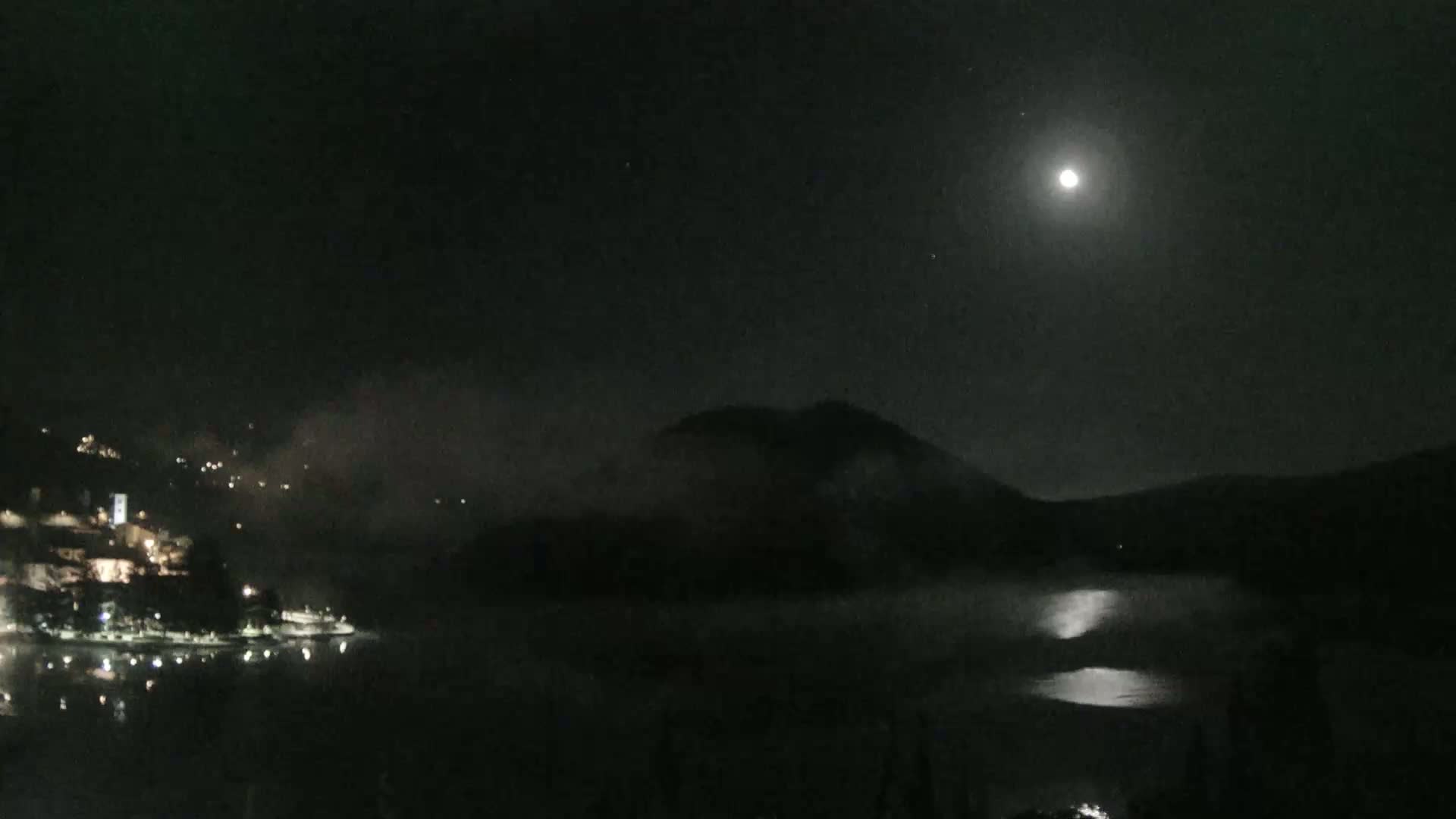 A moonlit night scene shows a body of water with a town's lights visible on one shore and a hill shrouded in fog on the opposite side.