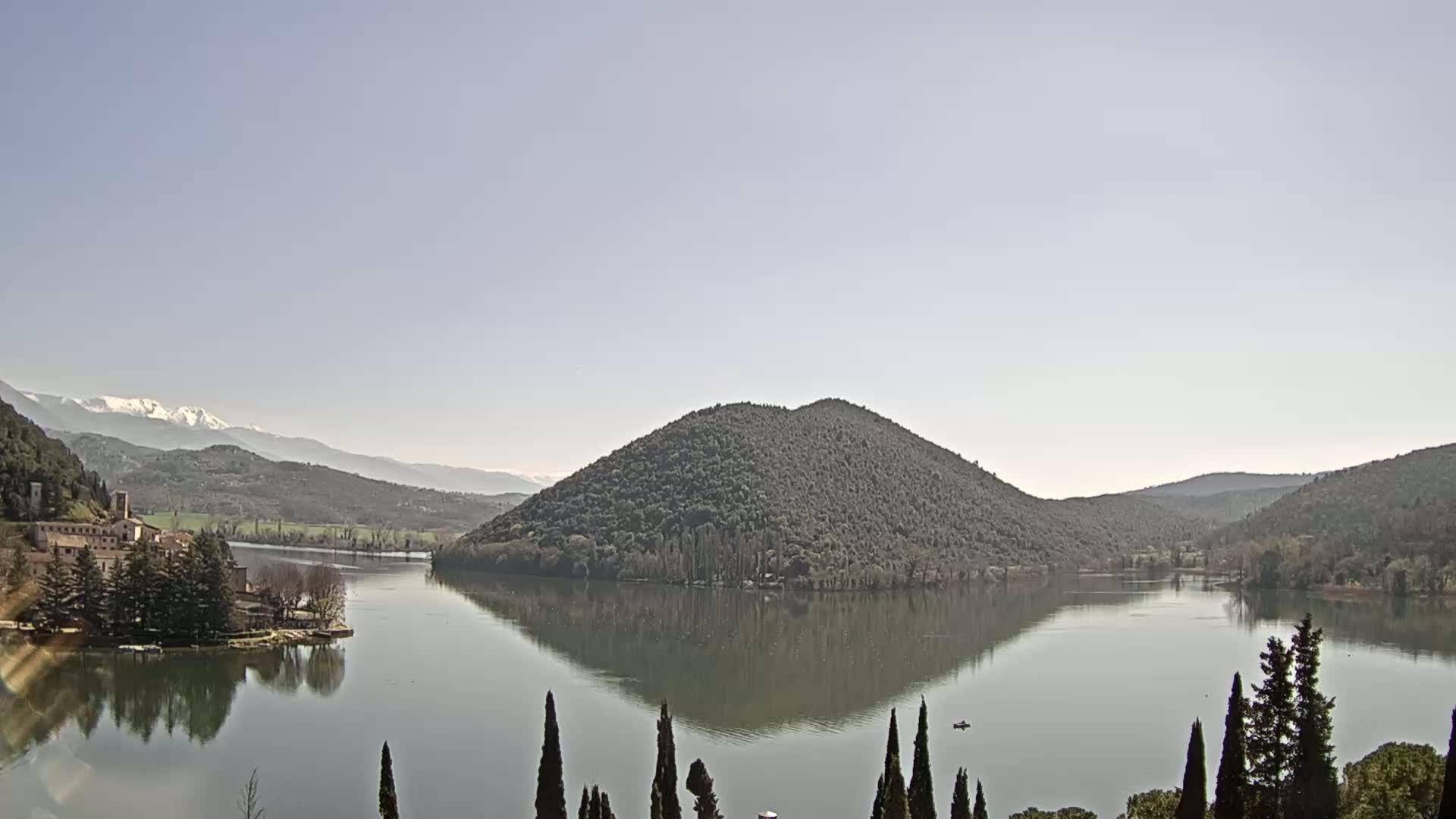 A calm lake reflects a tree-covered hill and snow-capped mountains under a clear sky.