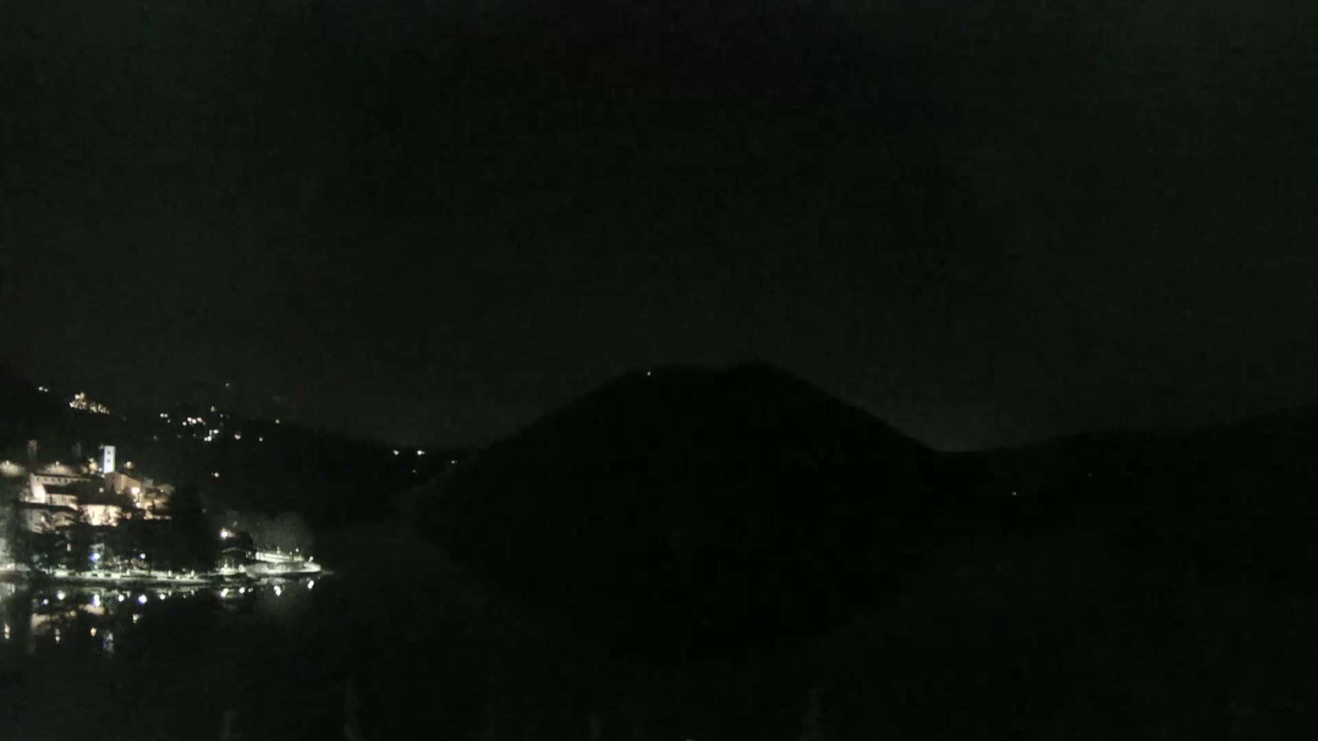 A nighttime view of a lake, with a dark hill in the background and a town with lights visible on the opposite shore.