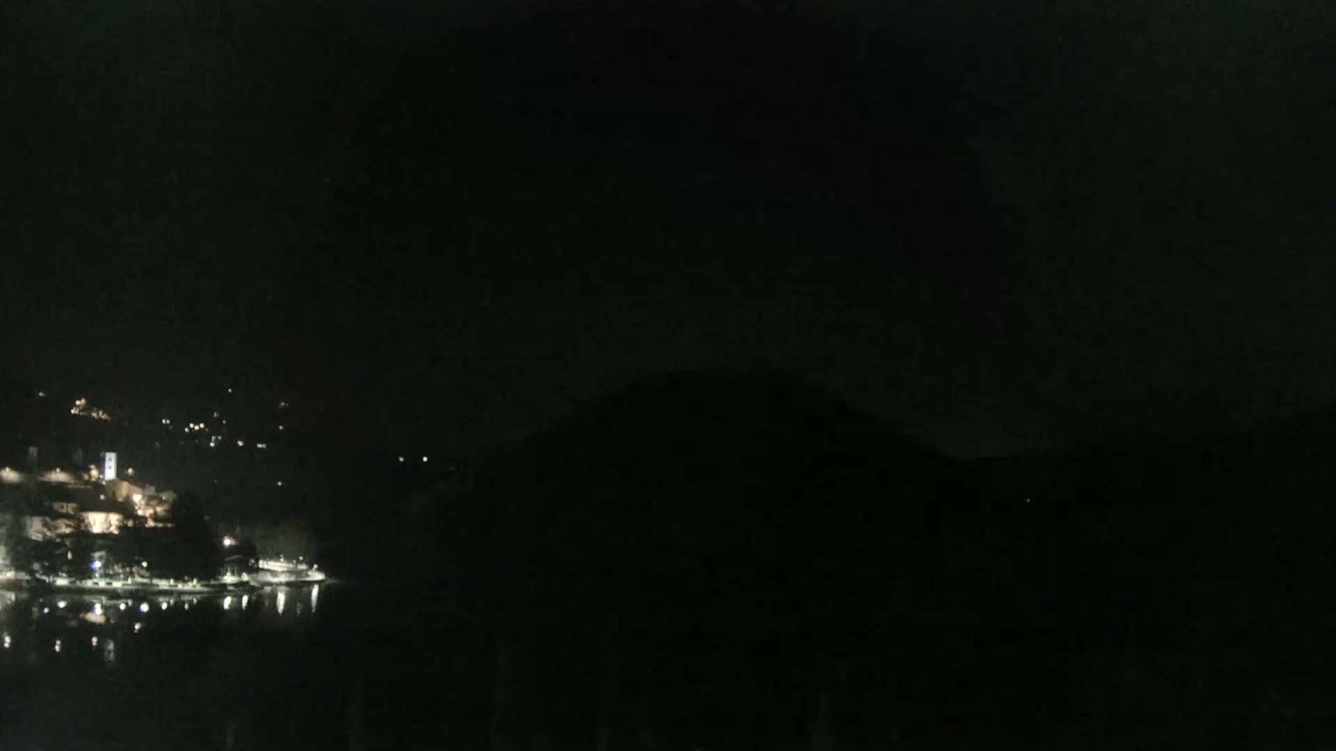 A nighttime view of a dimly lit town situated on a waterfront, with dark hills in the background under a clear, dark sky.