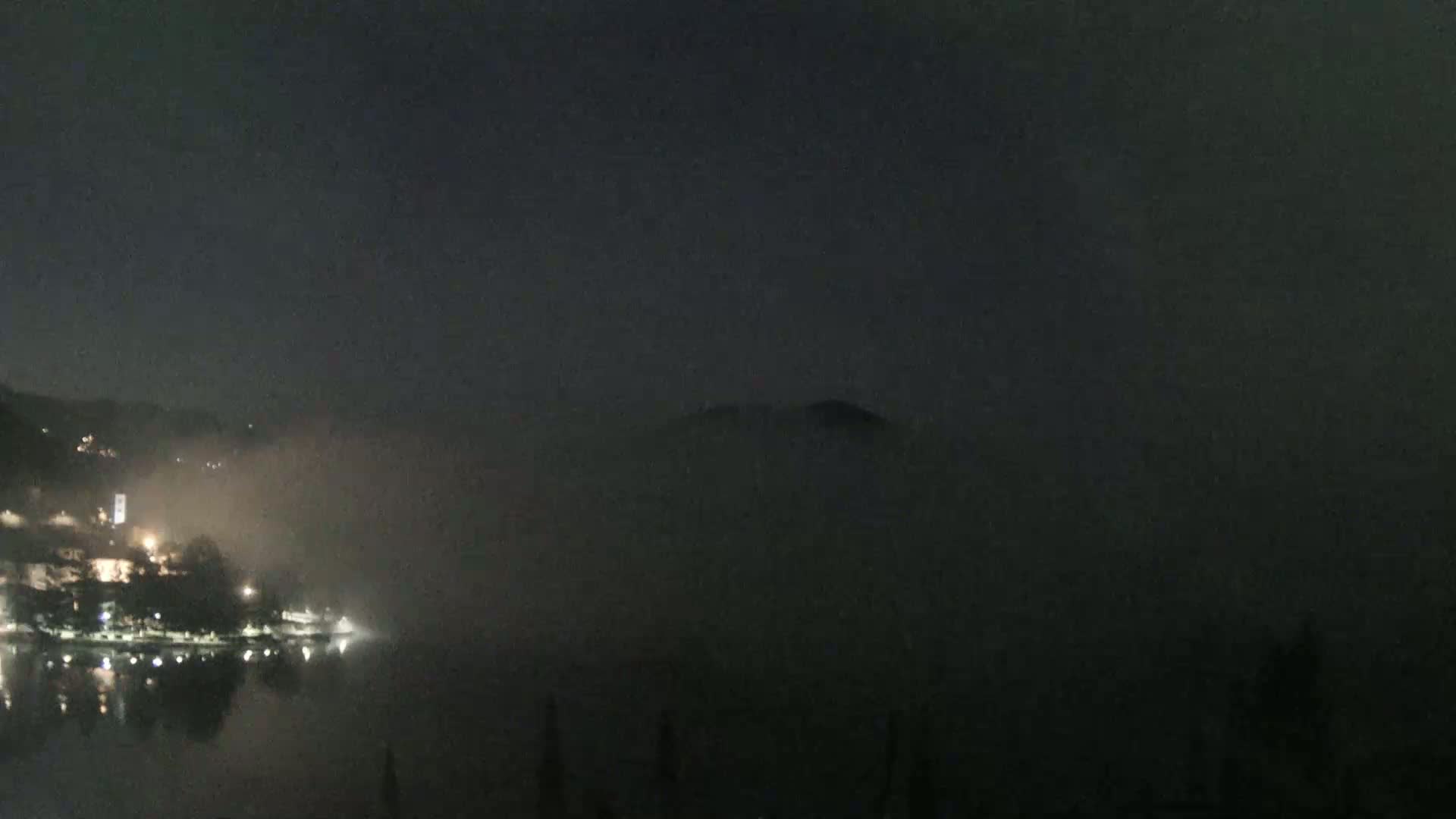 A nighttime view of a town on a lake shrouded in fog, with dimly lit buildings and a dark, hazy mountain in the background.