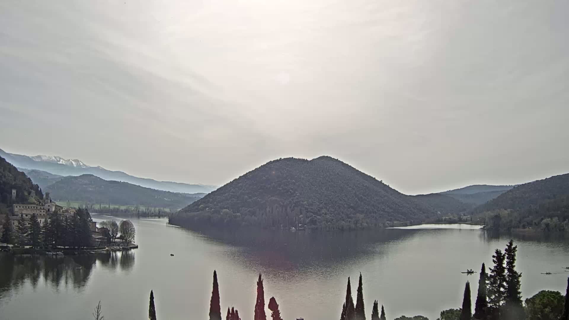 A calm lake surrounded by hills and mountains under a hazy sky shows snow-capped peaks in the distance.