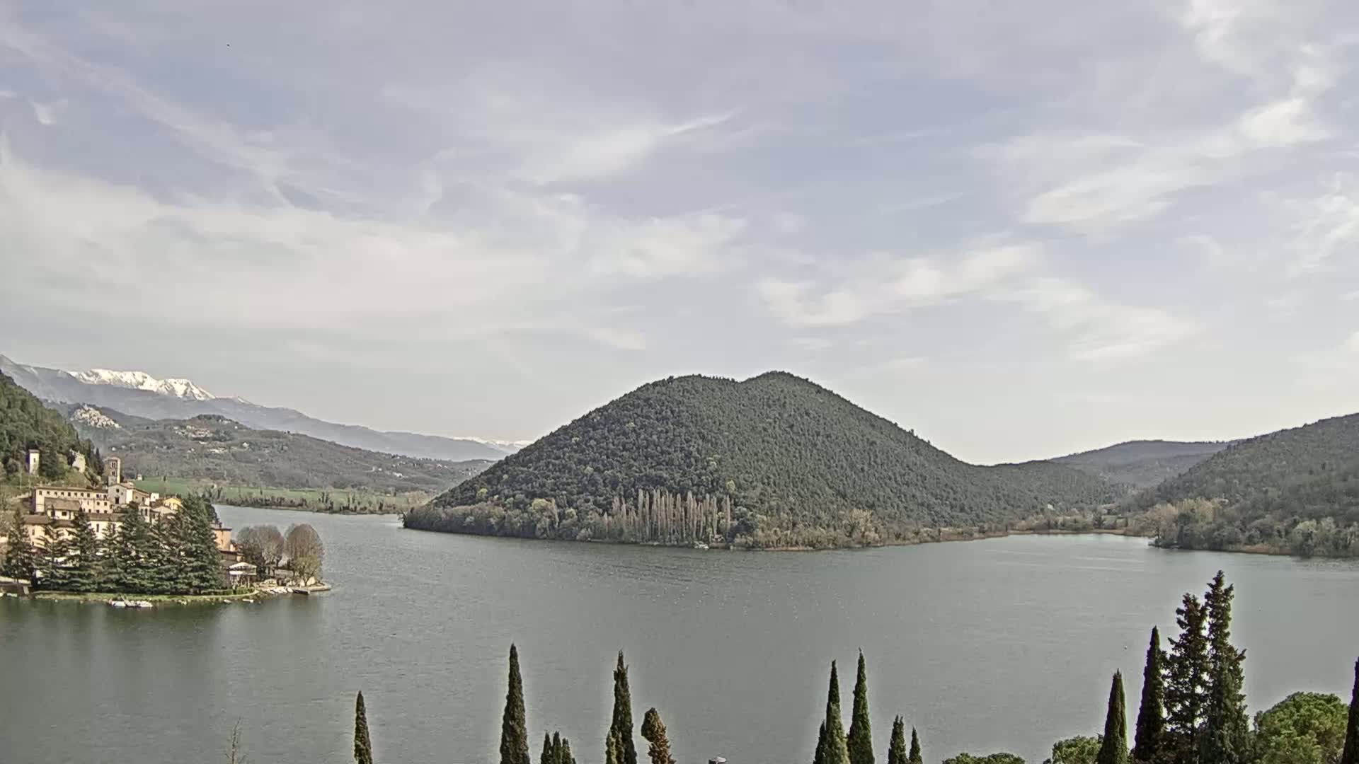 A lake is seen under a partly cloudy sky, with a town and snow-capped mountains in the distance, and a large, dark green island in the middle of the lake.