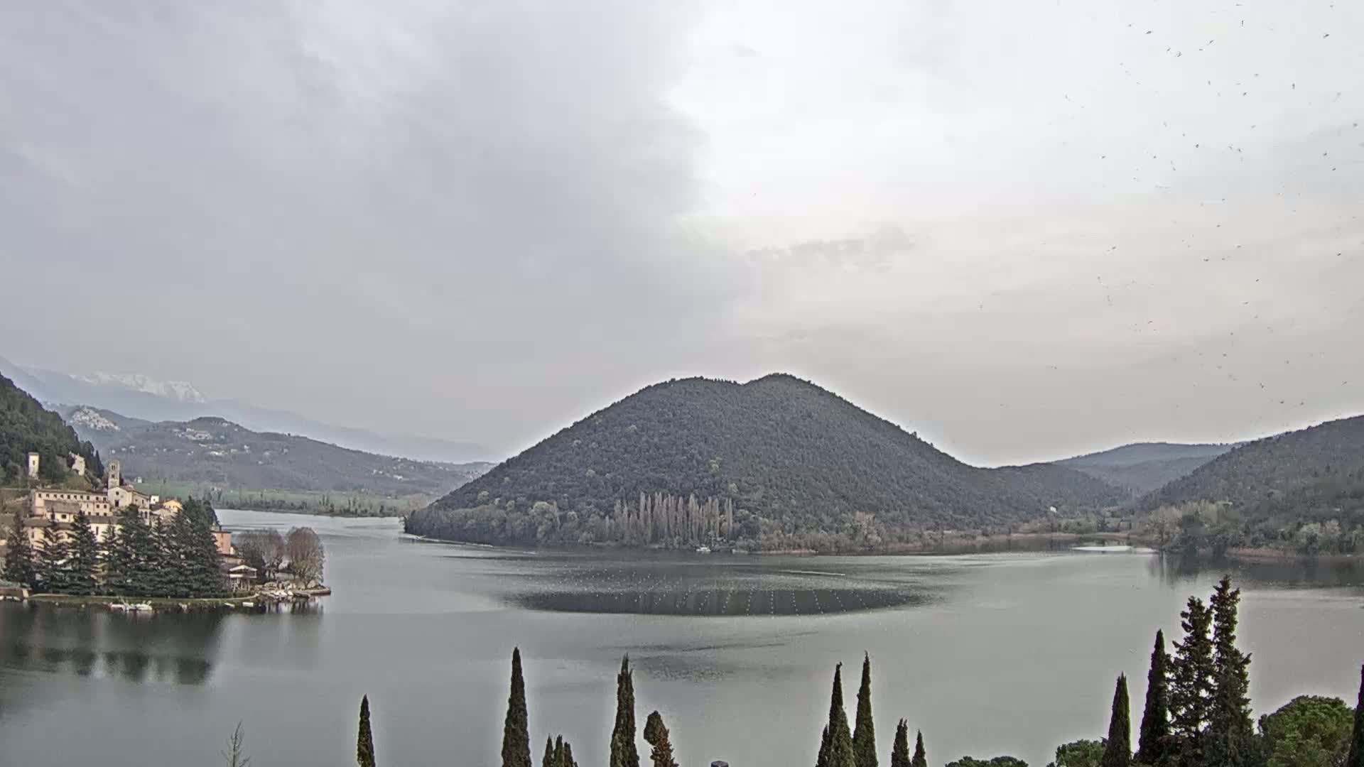 A lake is surrounded by hills and mountains under a cloudy sky, with a village nestled on its shore and a flock of birds flying overhead.