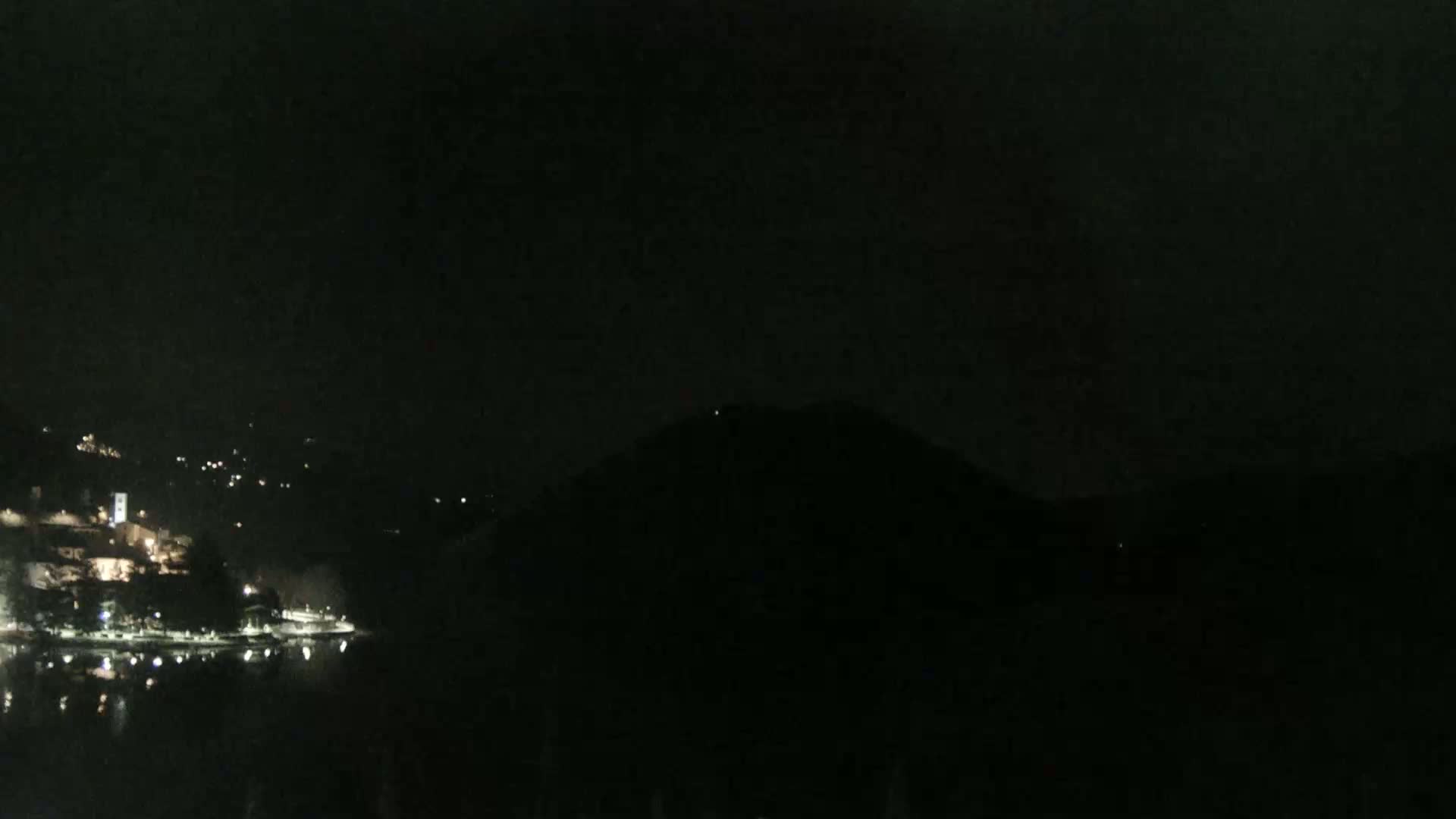 A dark nighttime scene shows a body of water reflecting scattered lights from a town nestled on a hillside, with a larger, dark hill or mountain dominating the background under a mostly clear, dark sky.