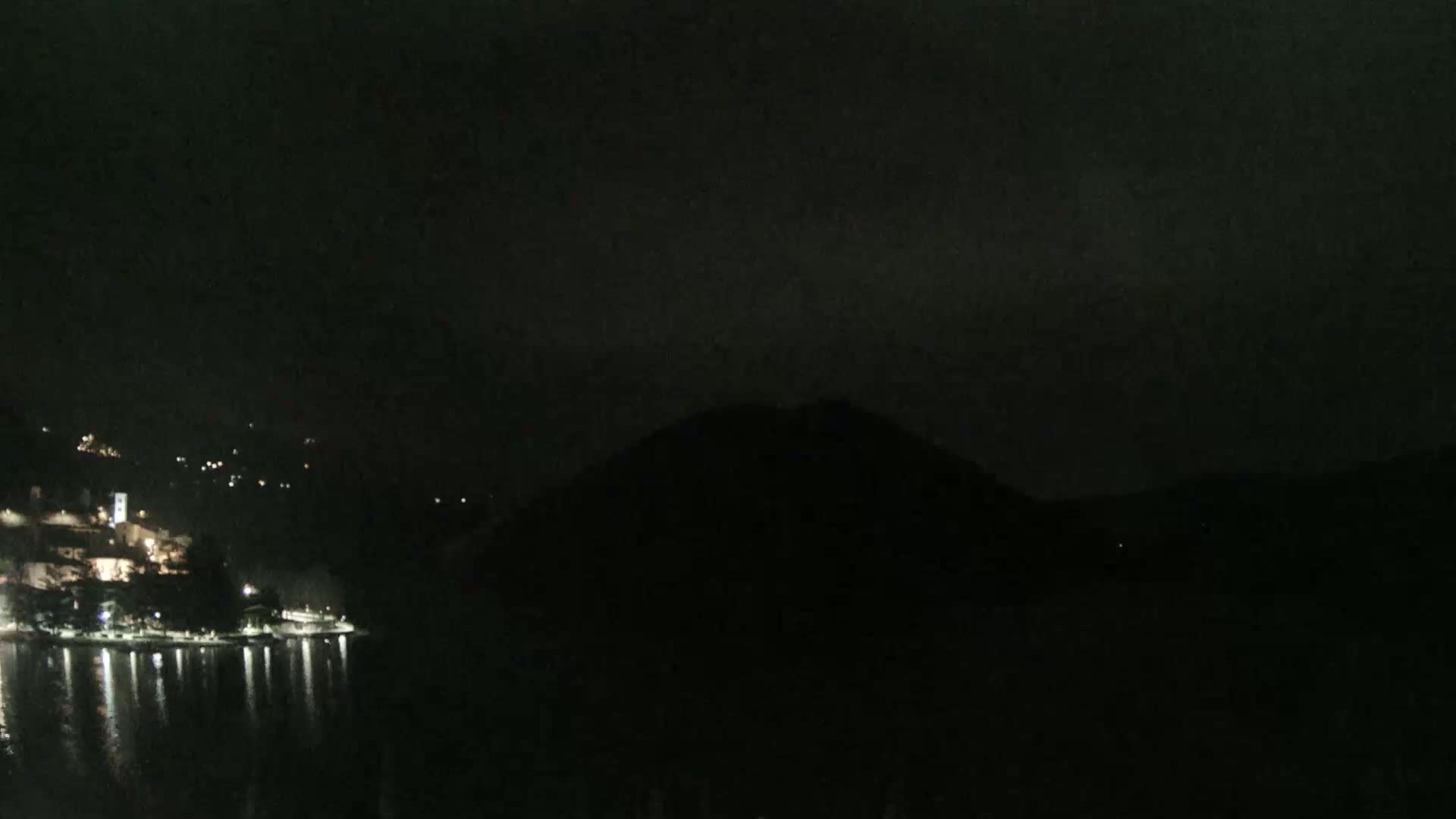 A dark, nighttime scene shows a body of water reflecting the lights of a small town nestled on a hillside, with a large, dark hill or mountain looming in the background under a mostly clear, dark sky.