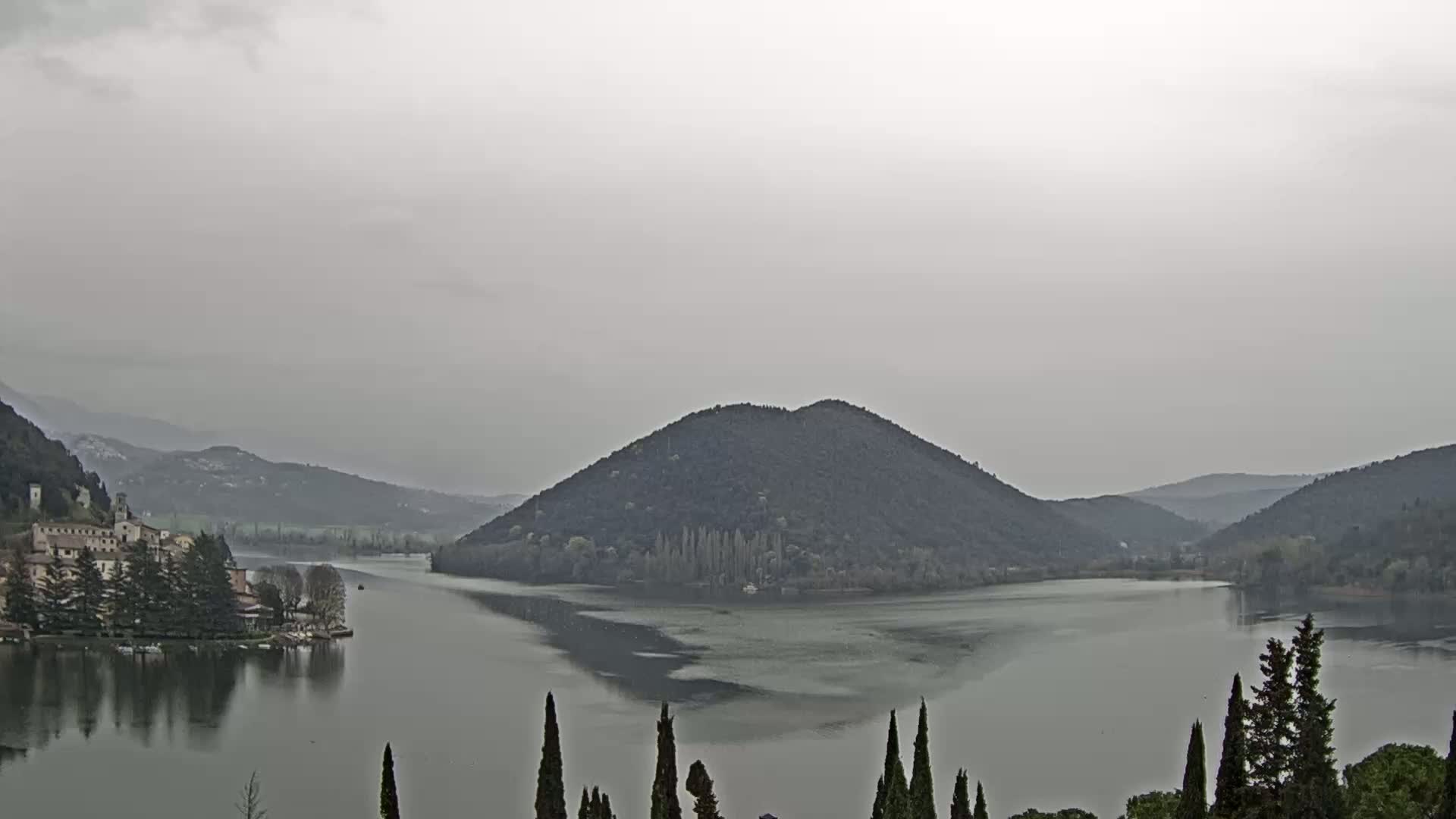 A calm lake surrounded by hills and trees under an overcast sky.
