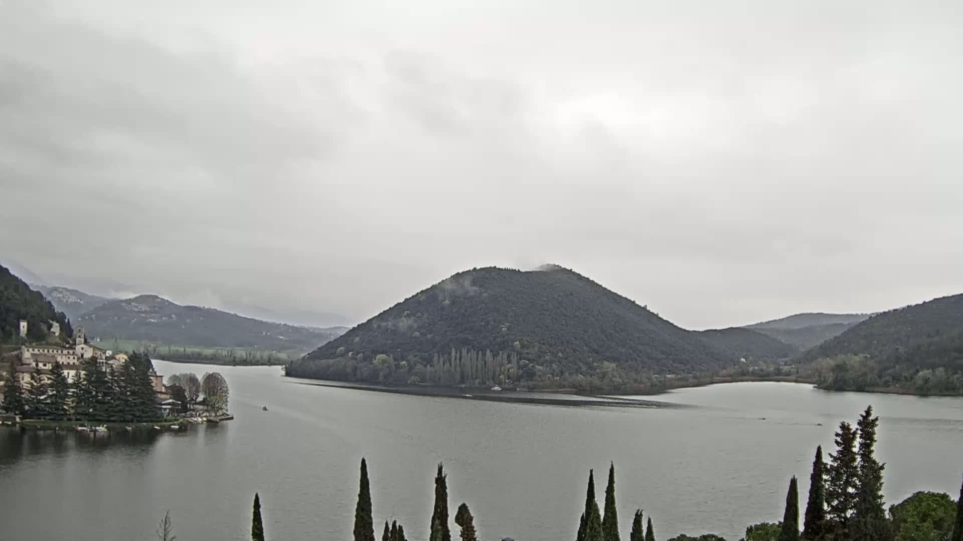 A lake is seen under an overcast sky, with a village and a tree-covered hill in the distance.