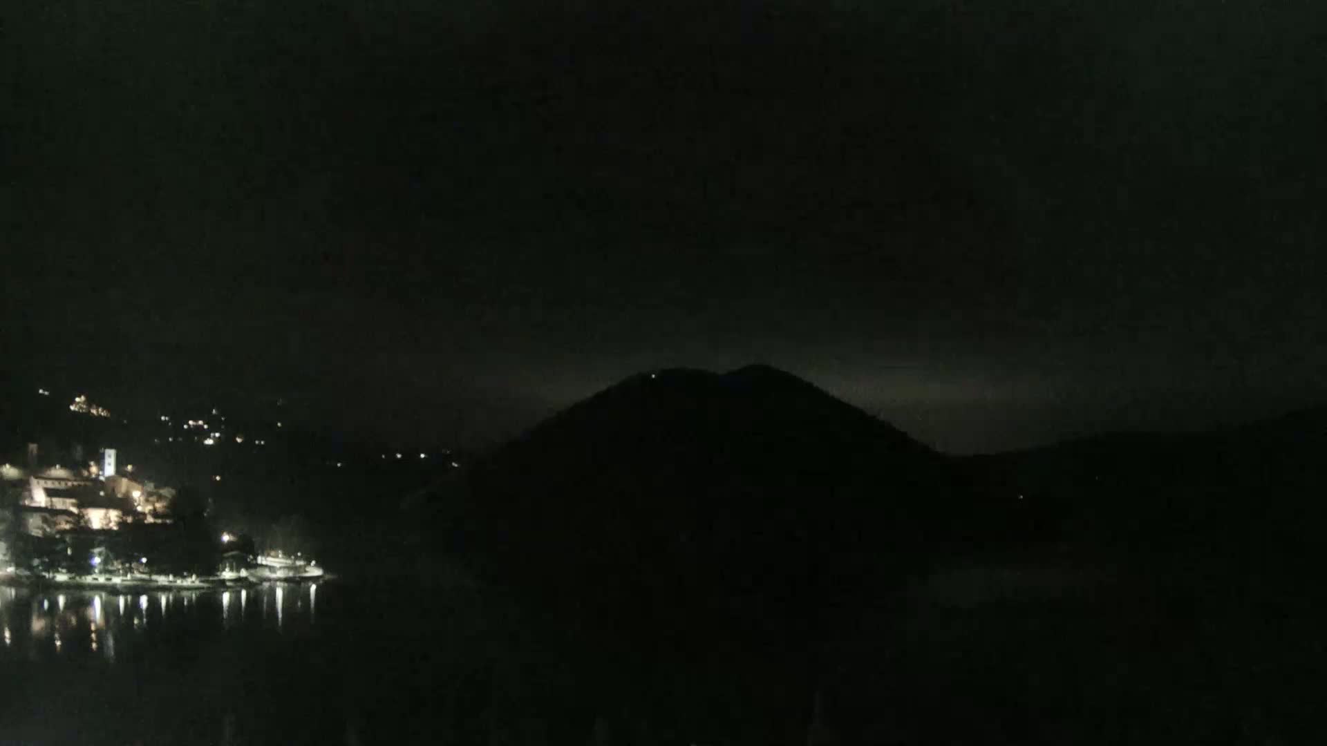 A nighttime view shows a dark hill silhouetted against a cloudy sky, with a small town's lights reflected on a calm body of water in the foreground.