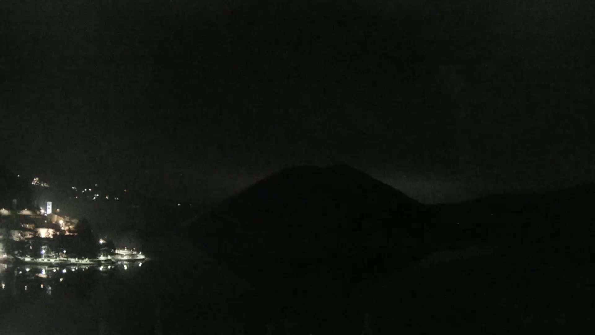 A nighttime view shows a dark, silhouetted hill overlooking a body of water with a small, lit-up town on the opposite shore.