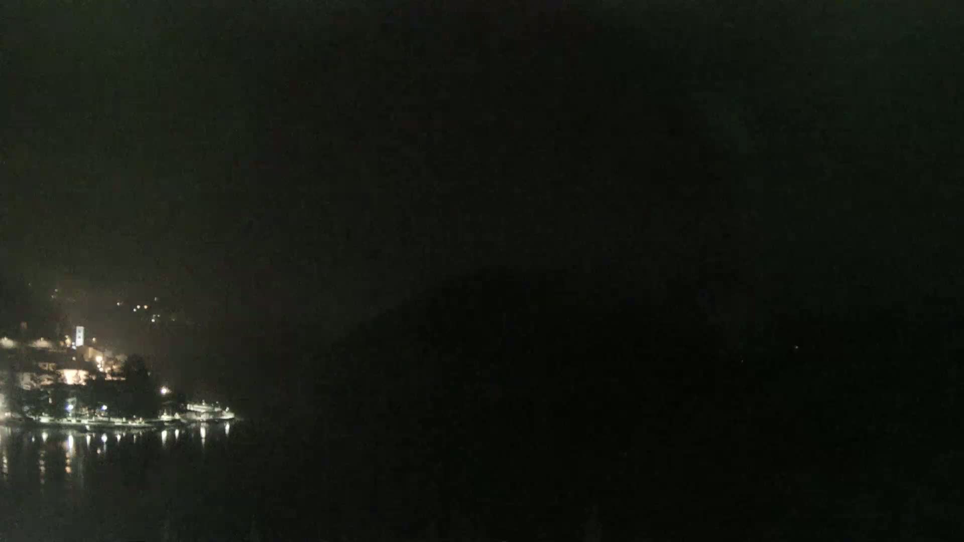 A nighttime, hazy view shows a dimly lit town situated along a dark body of water, with hills visible in the background.