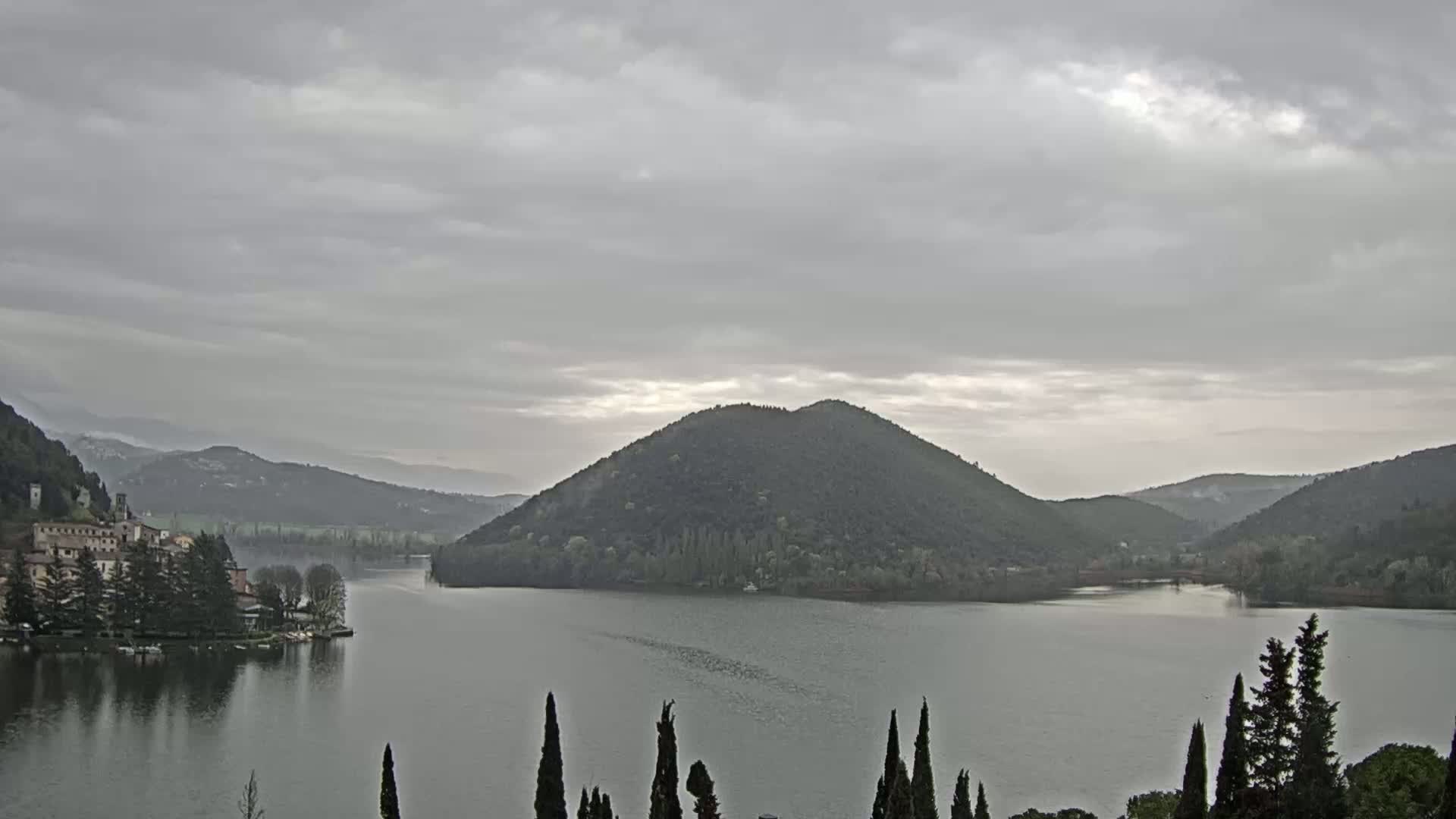 A lake is surrounded by rolling hills and a village under an overcast sky.