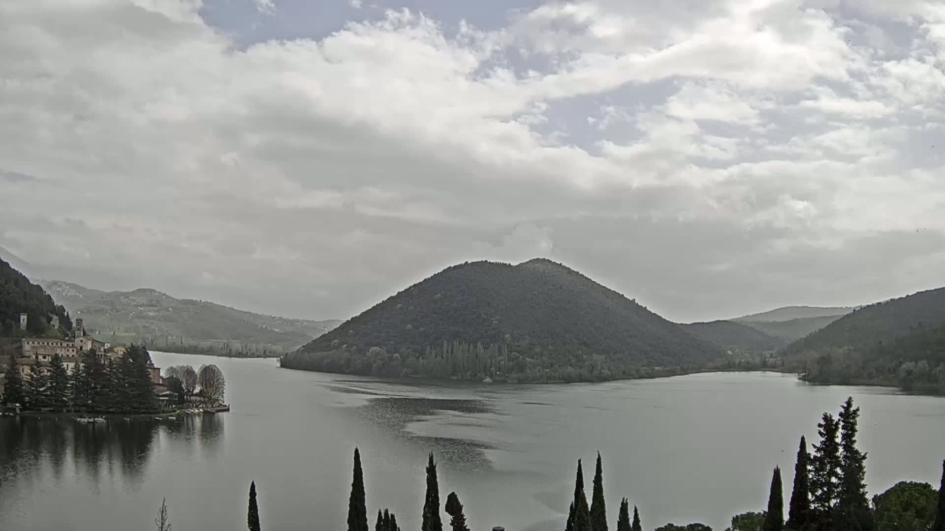 A lake is nestled between tree-covered hills under a mostly cloudy sky.