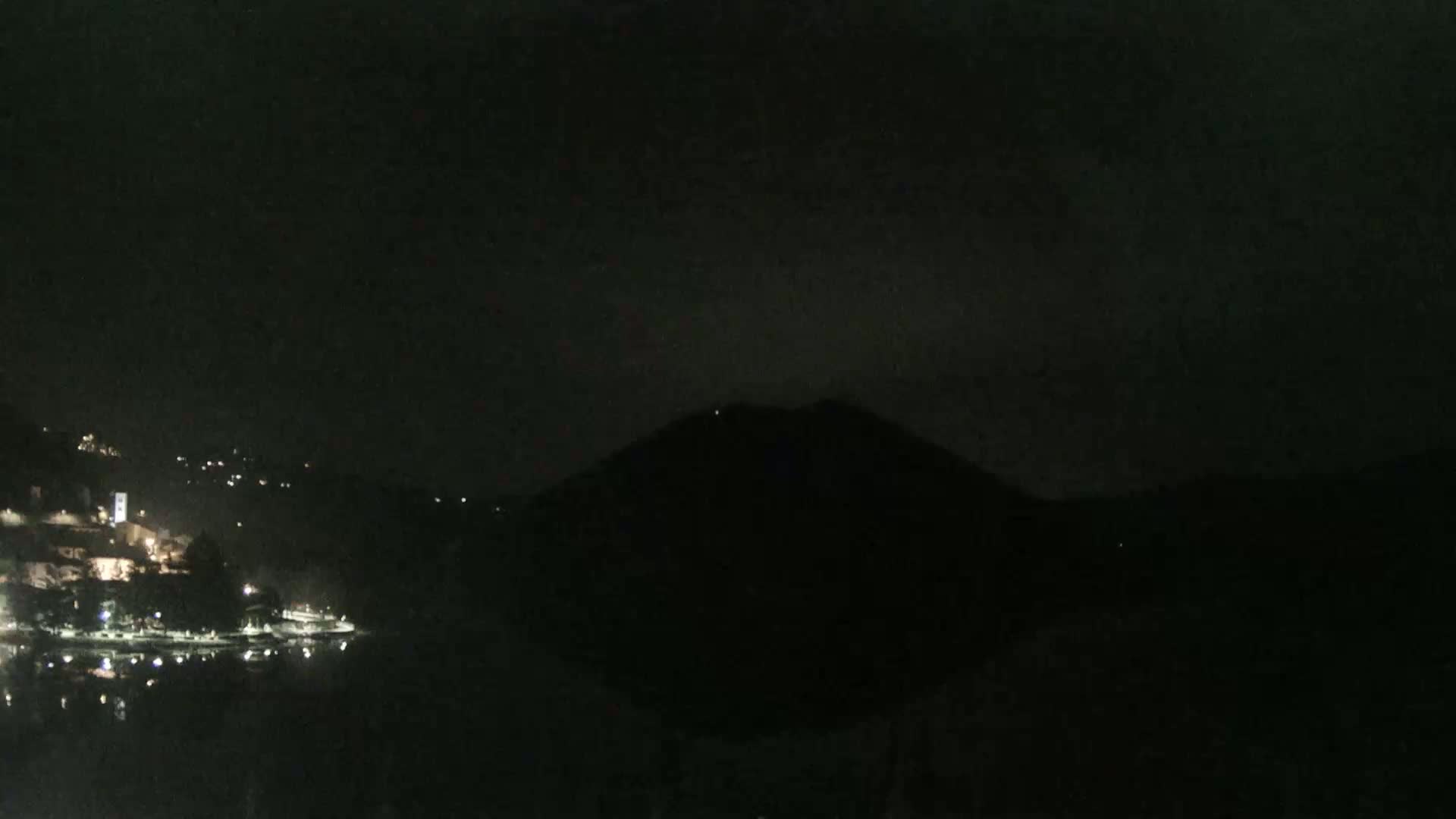 A nighttime view of a dark mountain overlooking a dimly lit lakeside town under a dark, cloudy sky.