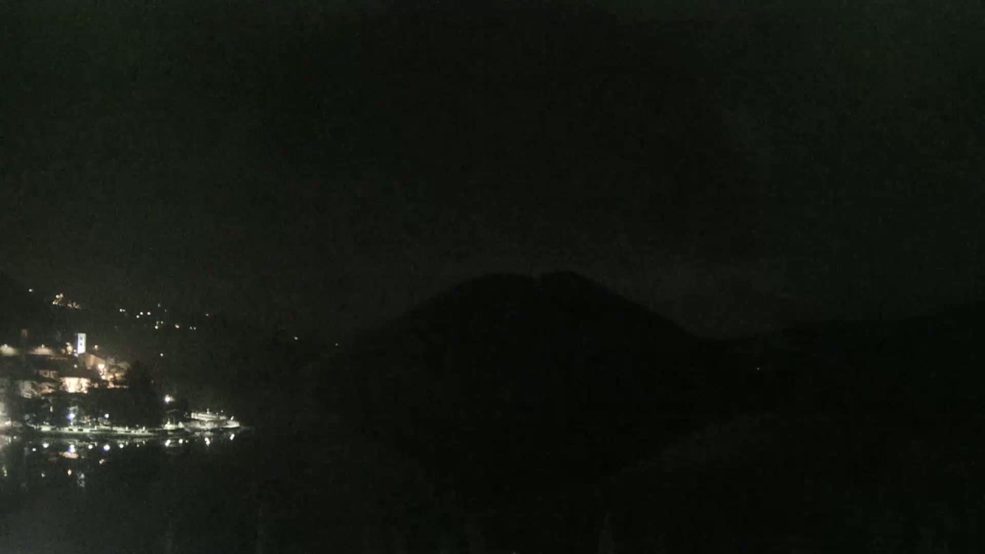 A nighttime view of a dimly lit town on a body of water, partially obscured by a dark hill under a clear, dark sky.