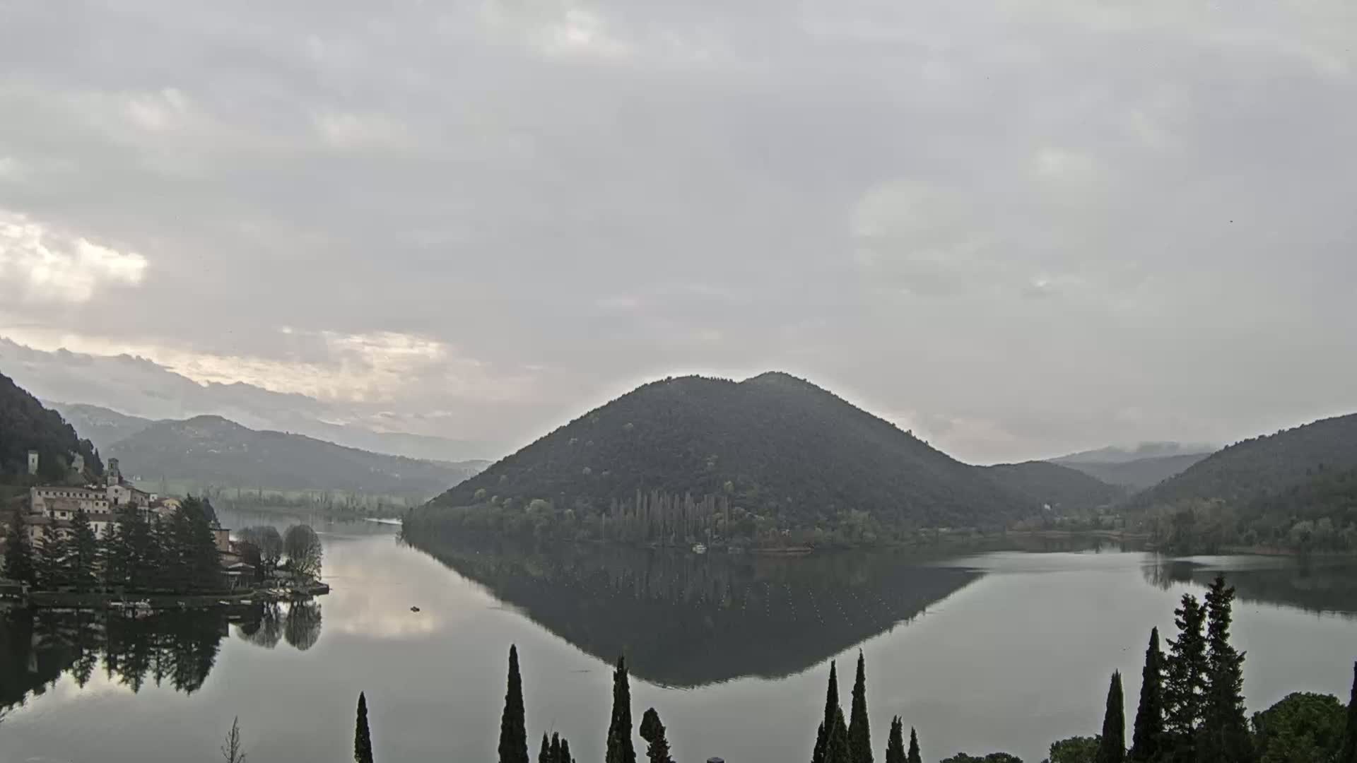 A calm lake reflects a tree-covered hill and a small village under an overcast sky.