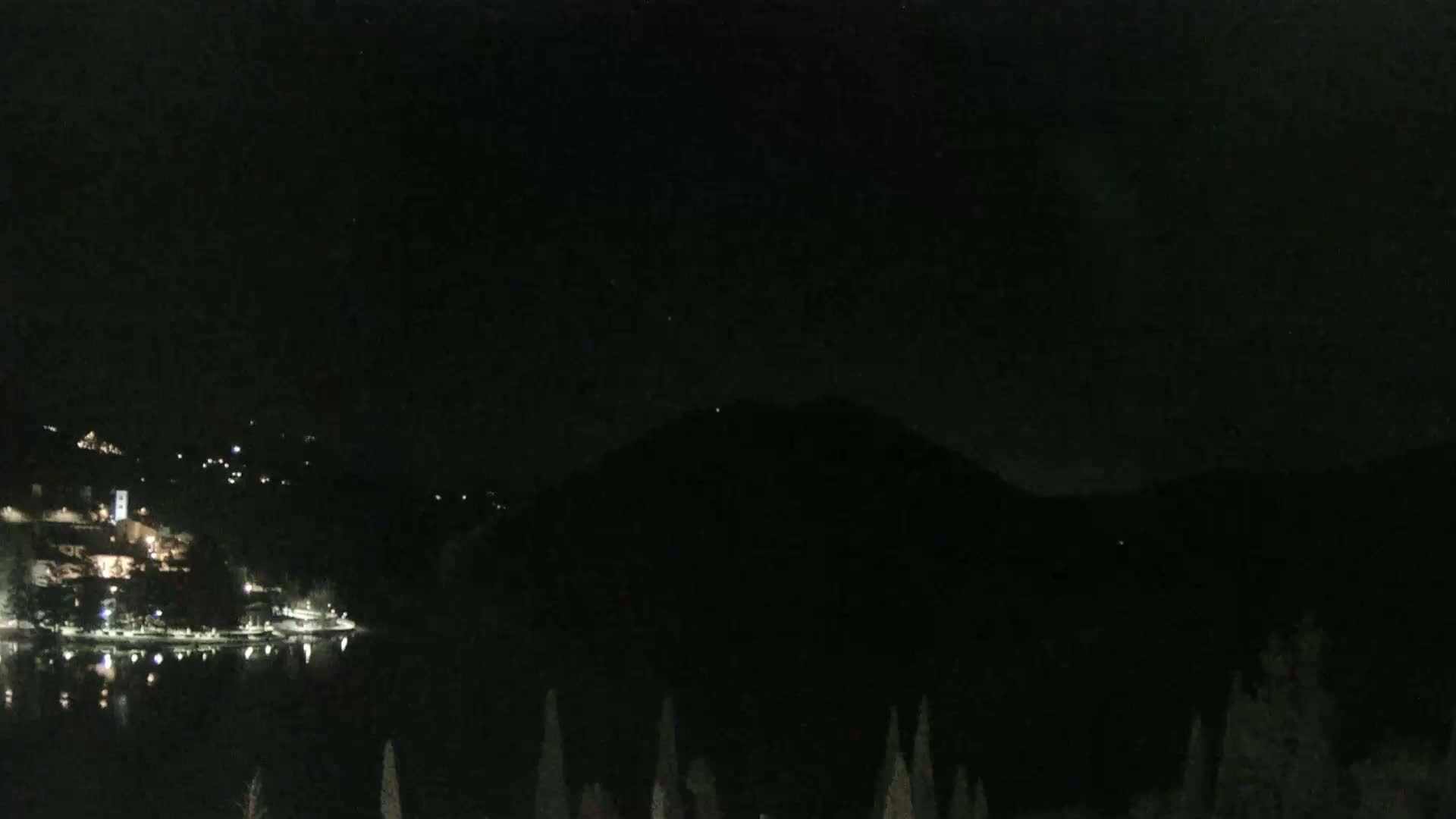 A nighttime view shows a dark lake reflecting a few lights from a town nestled on a hillside beneath a dark mountain.