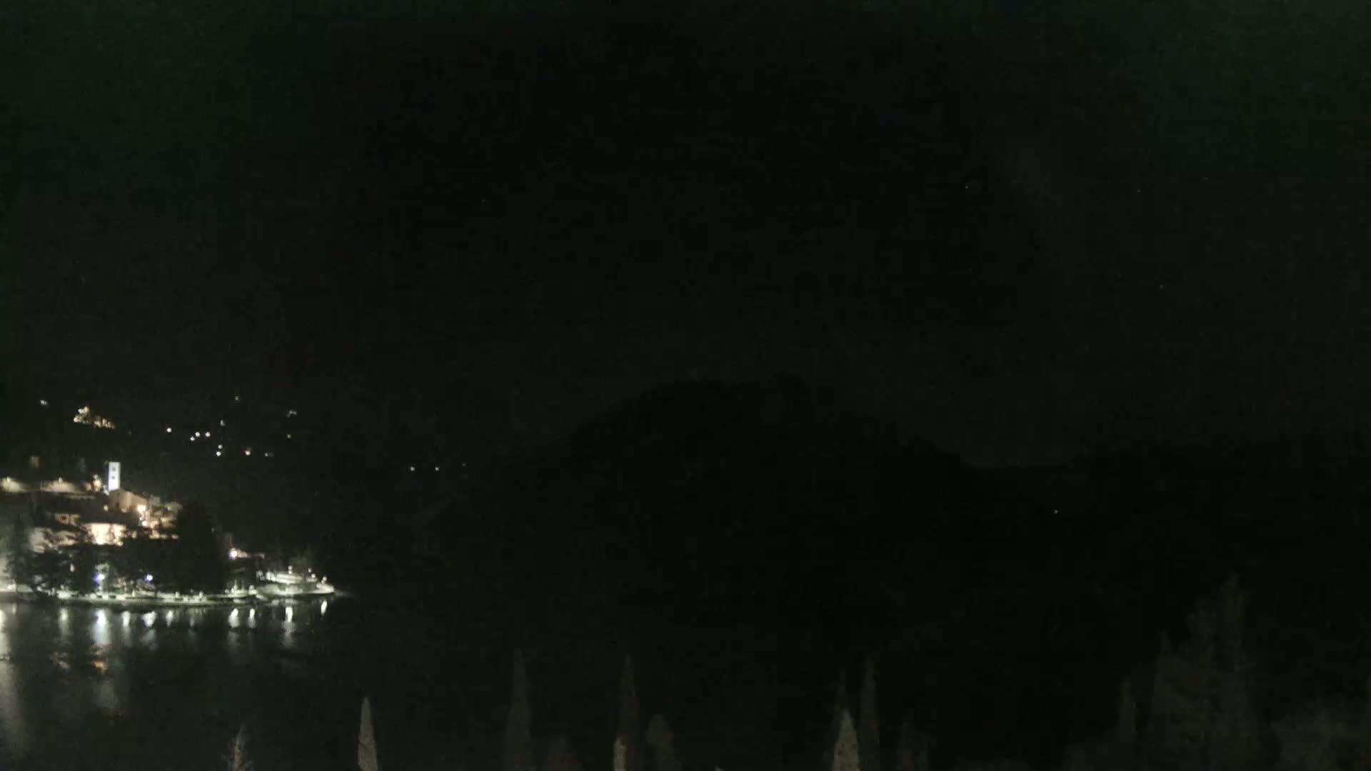 A dark nighttime scene shows a body of water reflecting the lights of a town nestled at the base of a hill.