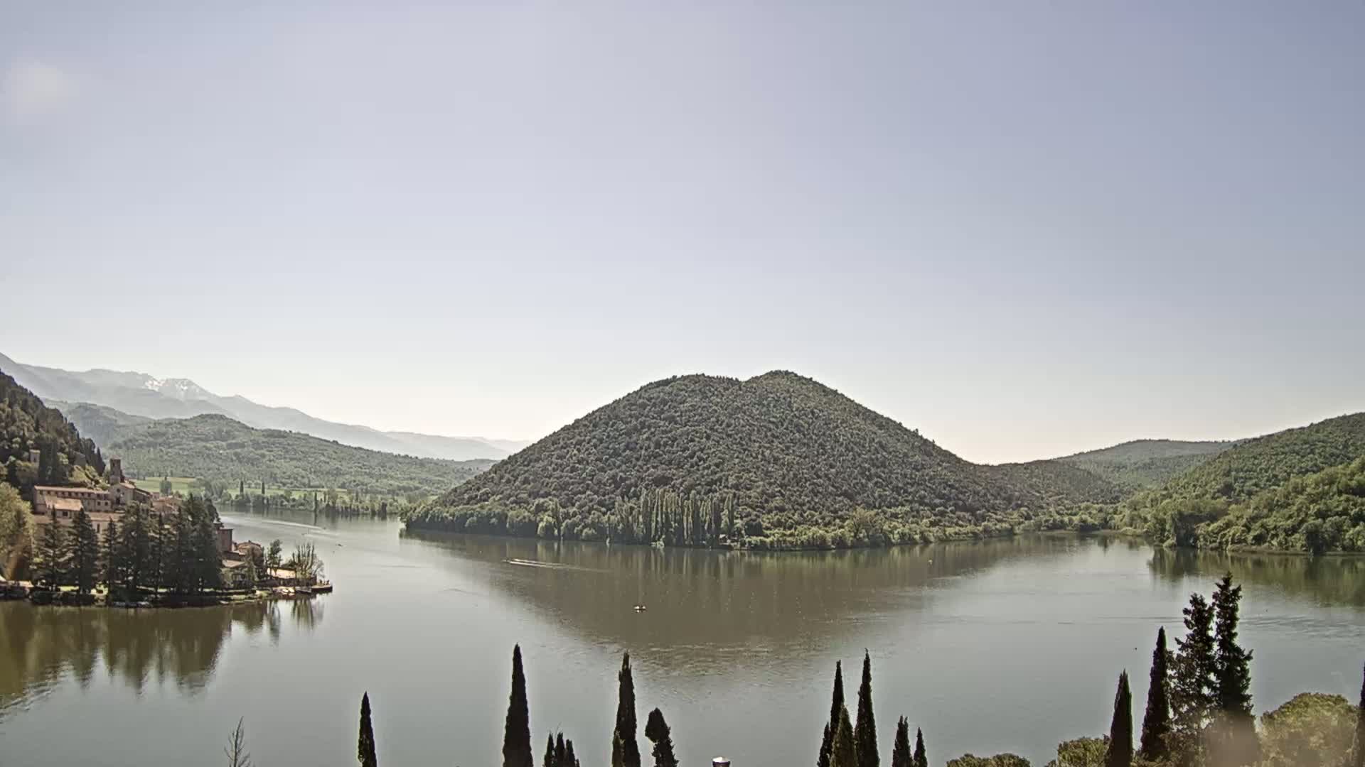 A calm lake is nestled between tree-covered hills and mountains under a clear, bright sky.