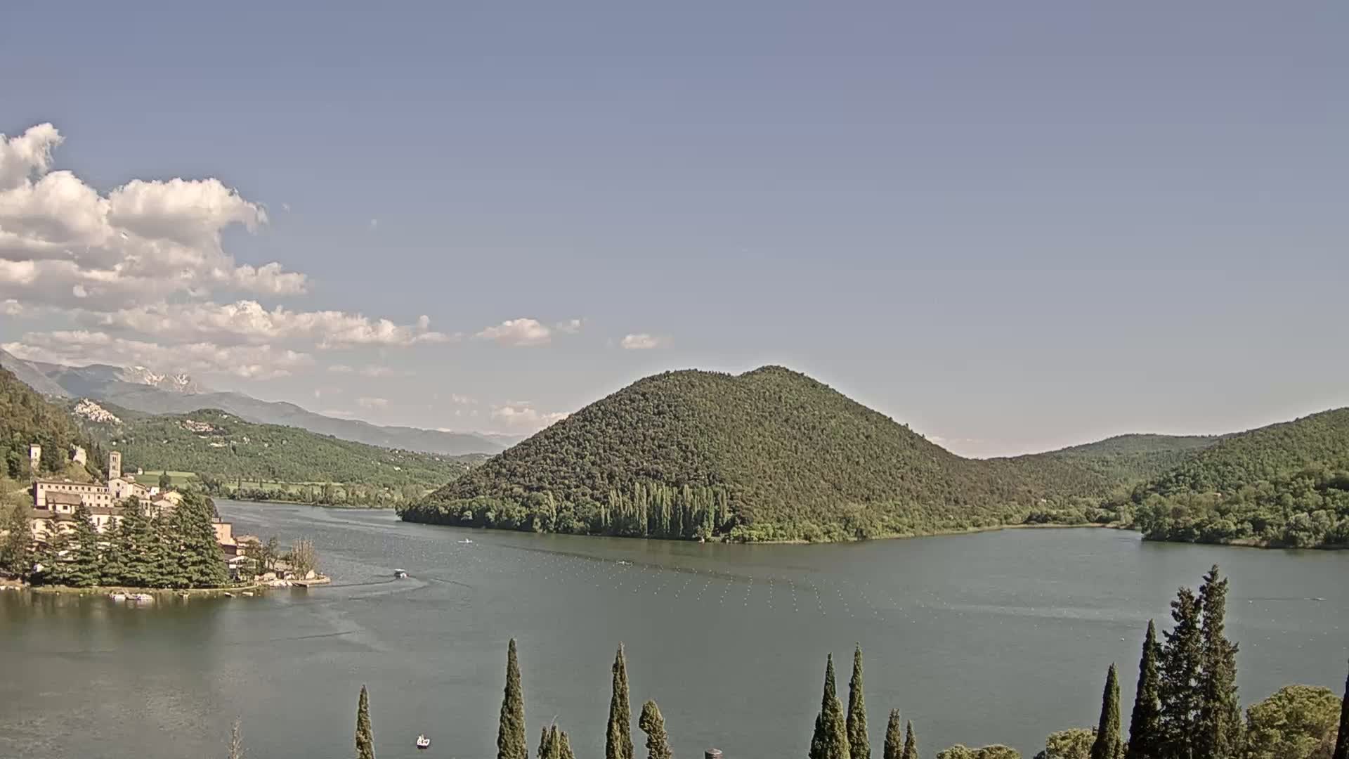 A tranquil lake is surrounded by lush green hills and mountains under a partly cloudy sky, with a small village nestled on the lakeshore.