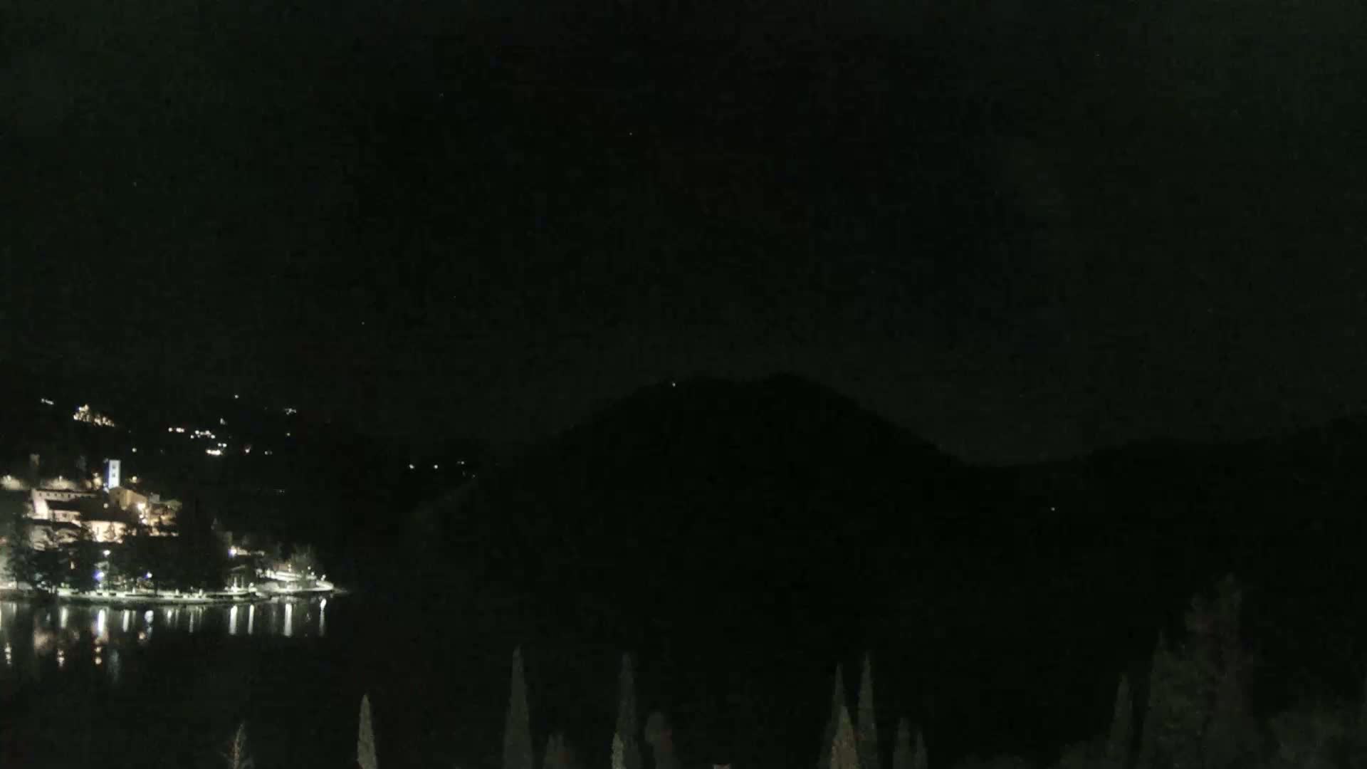 A nighttime view of a lake with a village and mountain in the background under a clear, dark sky.
