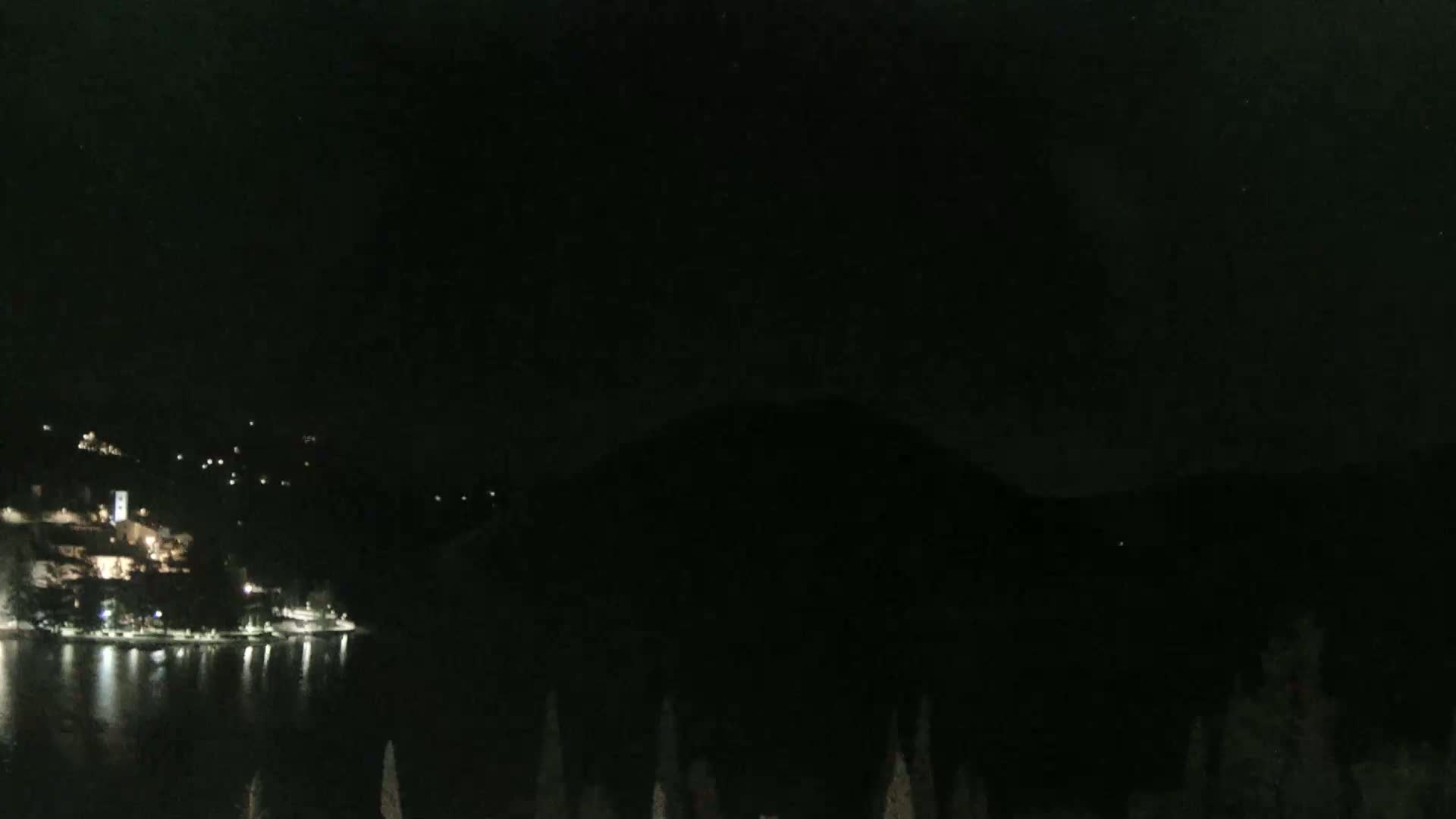 A dark nighttime scene shows a body of water reflecting the lights of a town on a hillside, with a dark mountain in the background.