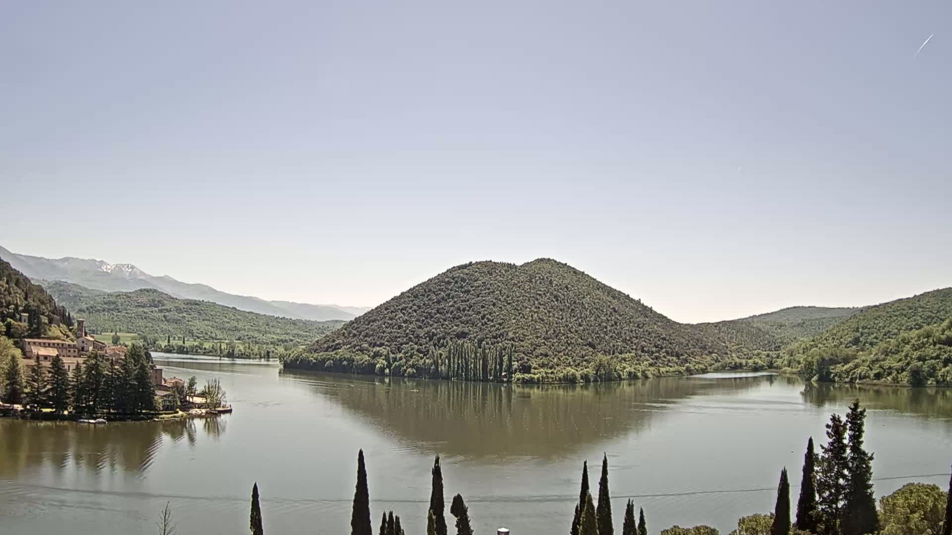 A calm lake nestled in a valley between green hills and mountains, under a clear, light-blue sky.