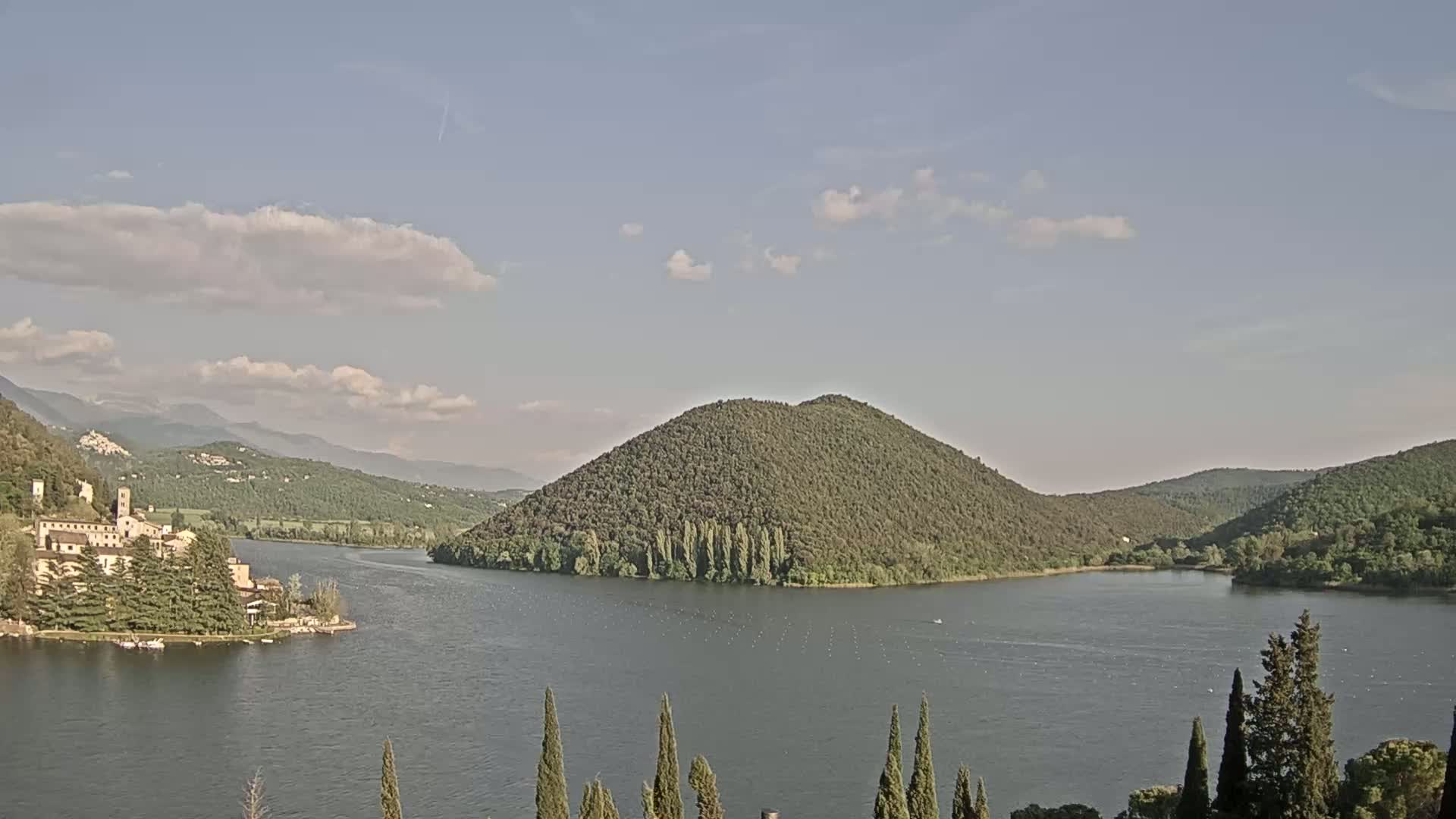 A lake with a large, tree-covered hill in the center, surrounded by other hills and mountains under a partly cloudy sky.